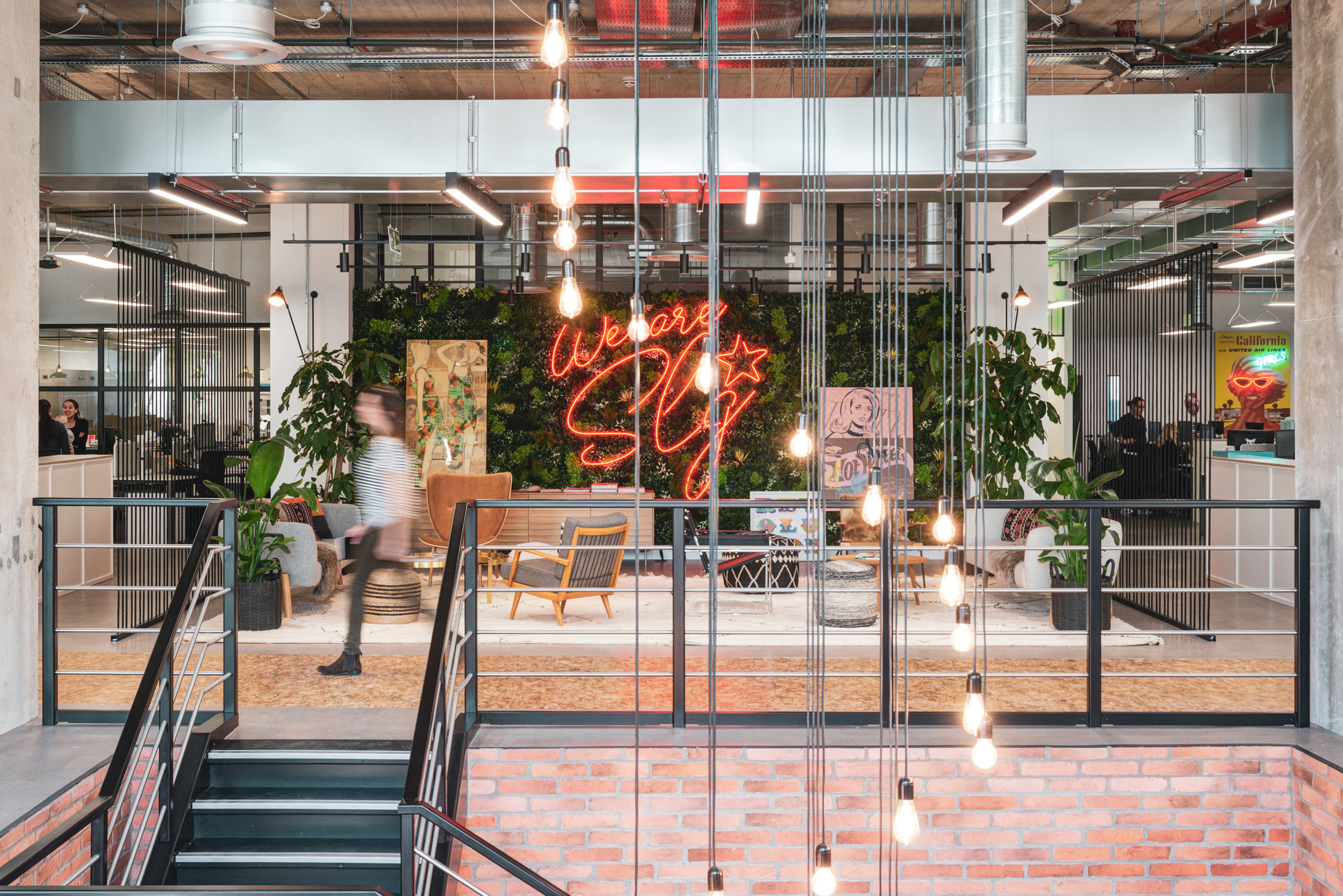 SLG’s vibrant award-winning workspace interior at the International Property Awards featuring exposed brick, hanging lights, and a neon sign reading 'We Are SLG,' with greenery and stylish seating areas in the background.