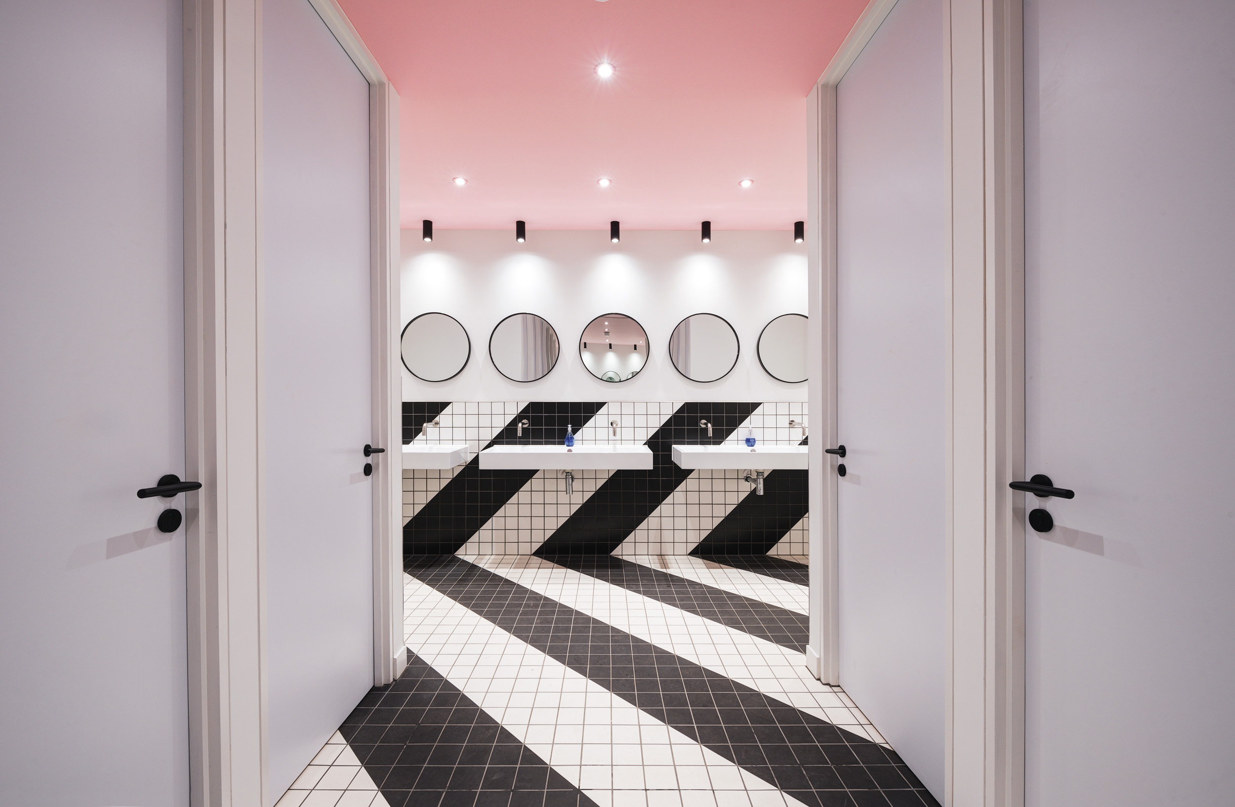 Studio 19’s stylish and contemporary award-winning bathroom interior design, featuring bold black and white geometric tile patterns, round mirrors, and a soft pink ceiling.