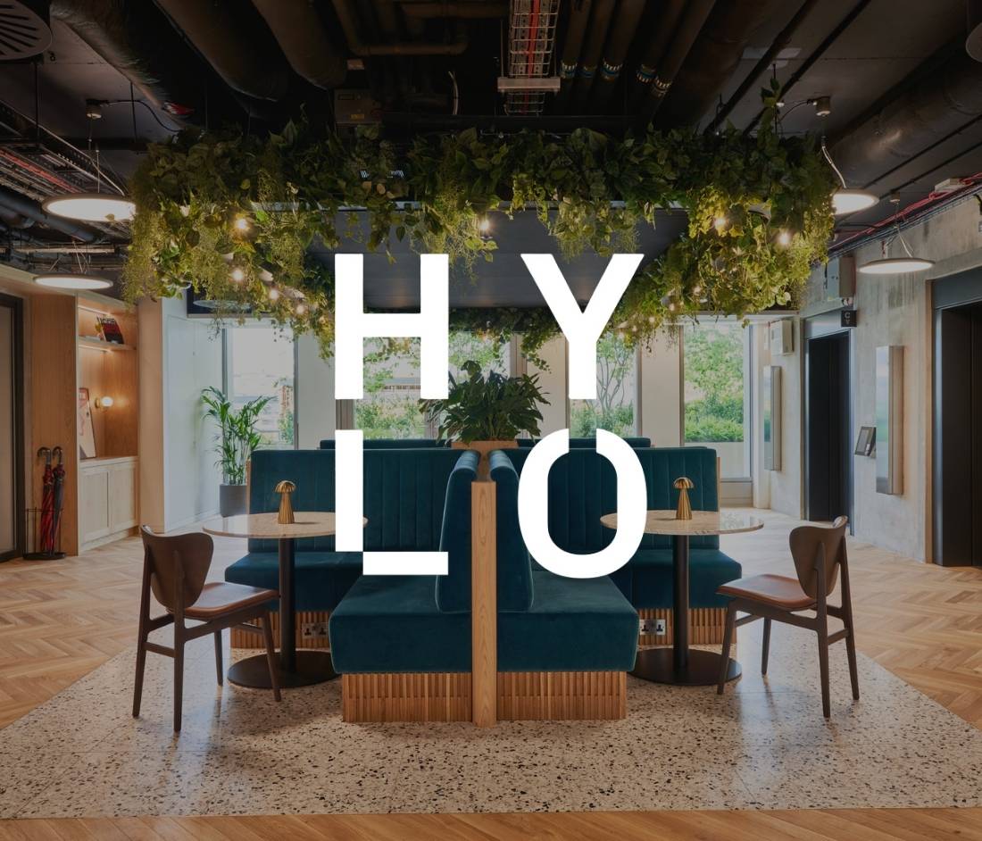 Modern office space in the HYLO building with teal booths, hanging greenery, and natural light.The word 'HYLO' is prominently displayed in bold, white letters in the foreground.