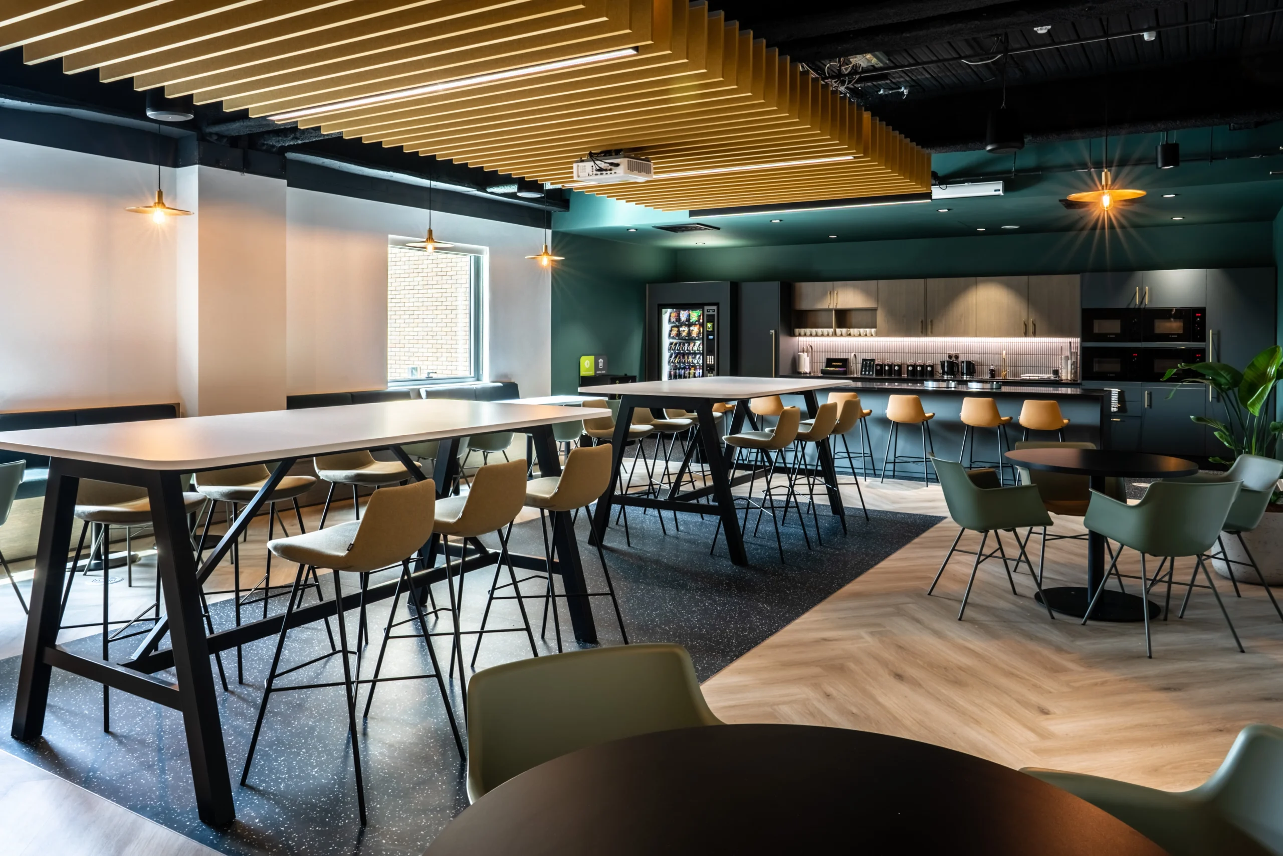 A contemporary teapoint area at the Watches of Switzerland Group headquarters featuring high-top tables, comfortable seating, and an equipped kitchen designed for employees to come together in a relaxed and moody setting.