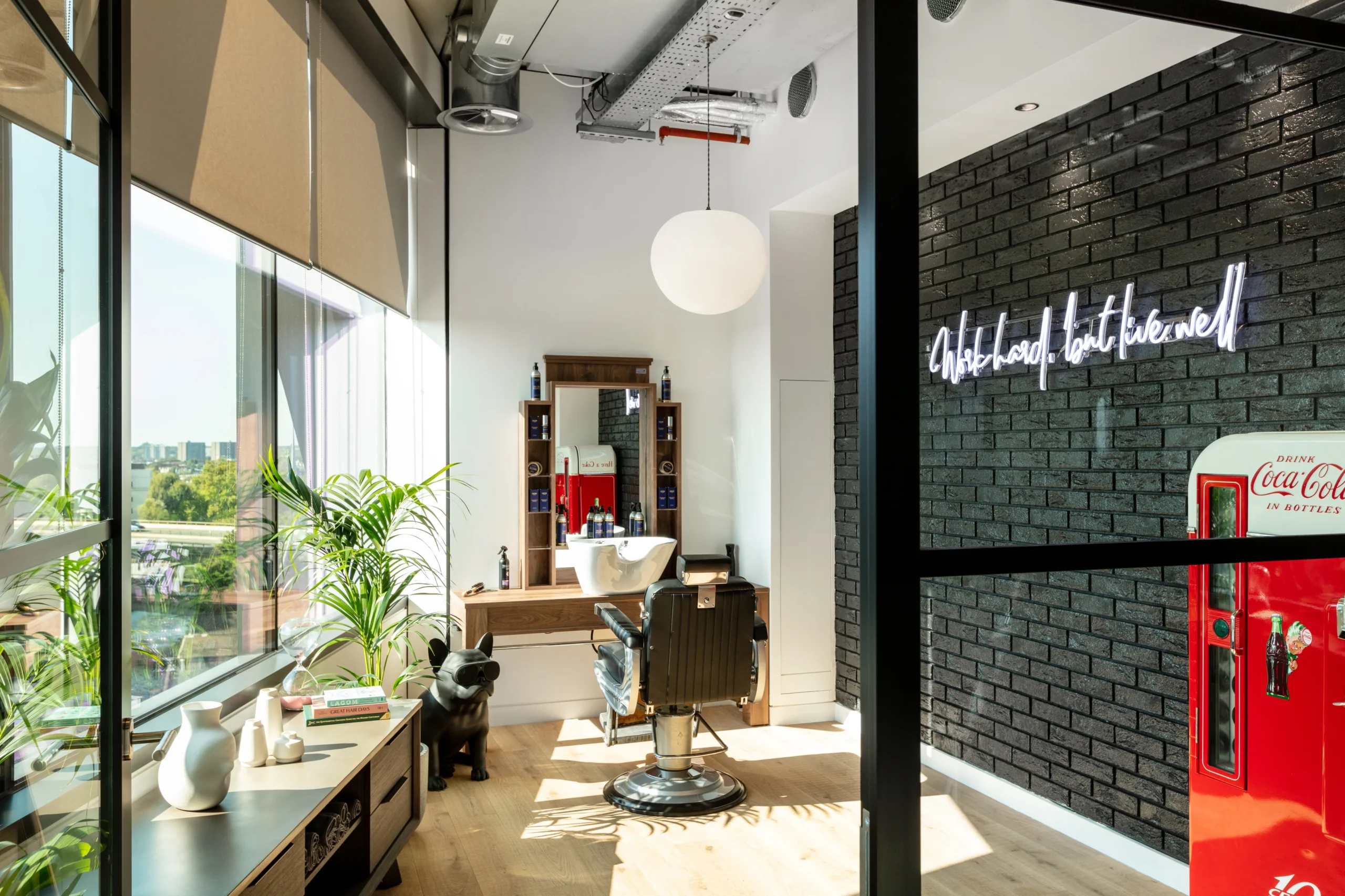 Alpha FX’s chic barber shop interior featuring a vintage-style barber chair, a wooden vanity with a mirror, and a bright red Coca-Cola fridge. A neon sign on a block wall reading, ‘Work Hard, Look Good’. Also, a few green plants and natural light.