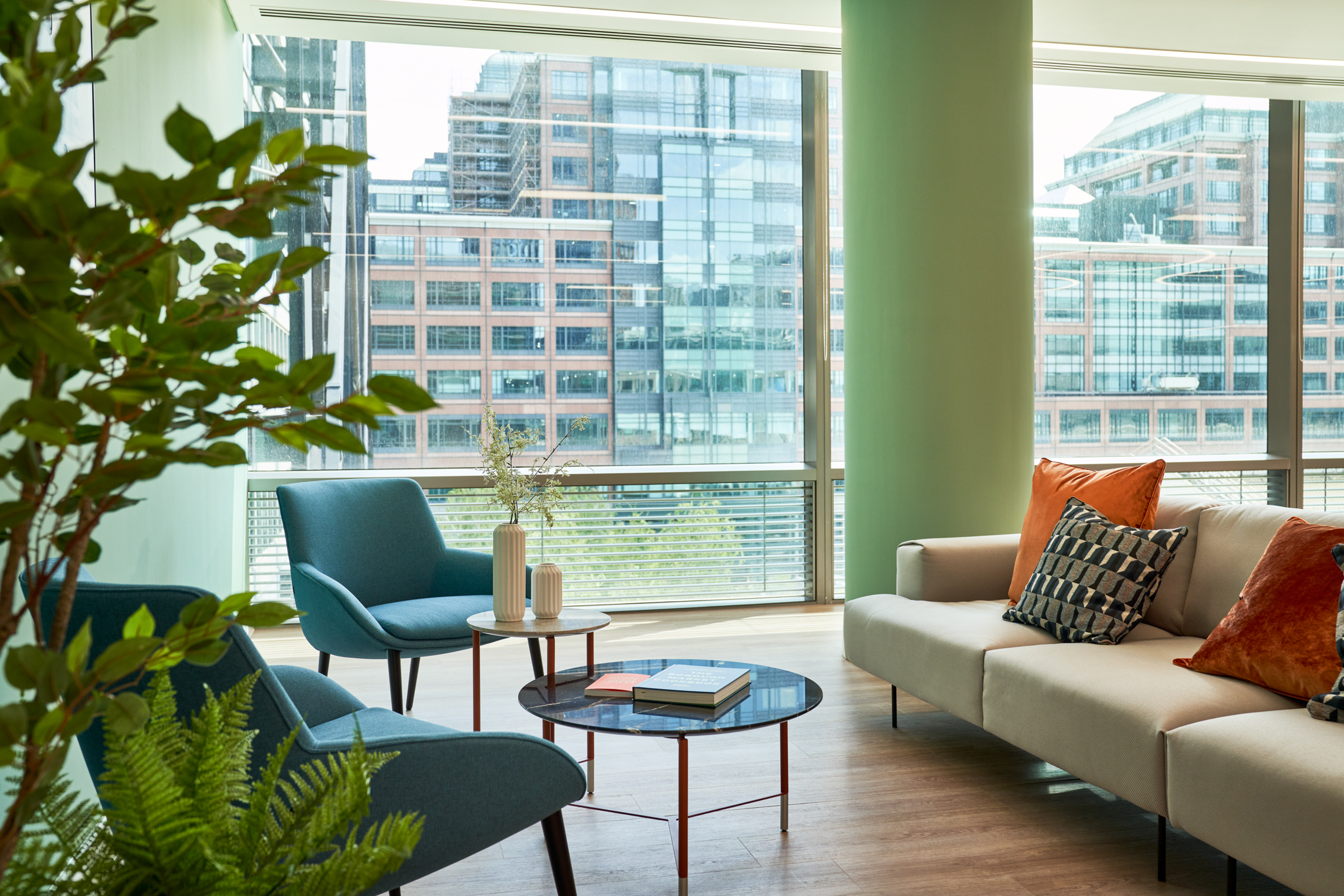 A cozy seating area in British Land's 10 Exchange Square, designed by Modus Workspace, featuring comfortable blue chairs and a beige sofa. The space is enhanced by large windows and vibrant green plants.