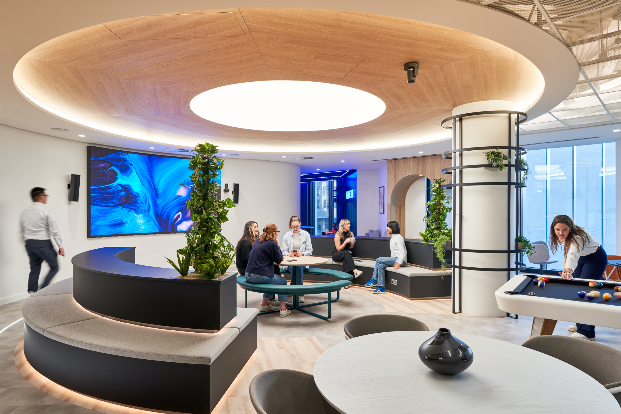 Capital.com office designed by Modus Workspace featuring a modern, open-plan communal area with curved seating, integrated planters, a large screen for digital display, and a pool table. The space emphasises collaboration with a mix of casual seating options and bright, natural lighting filtered through the circular ceiling feature, creating a contemporary and inviting atmosphere.