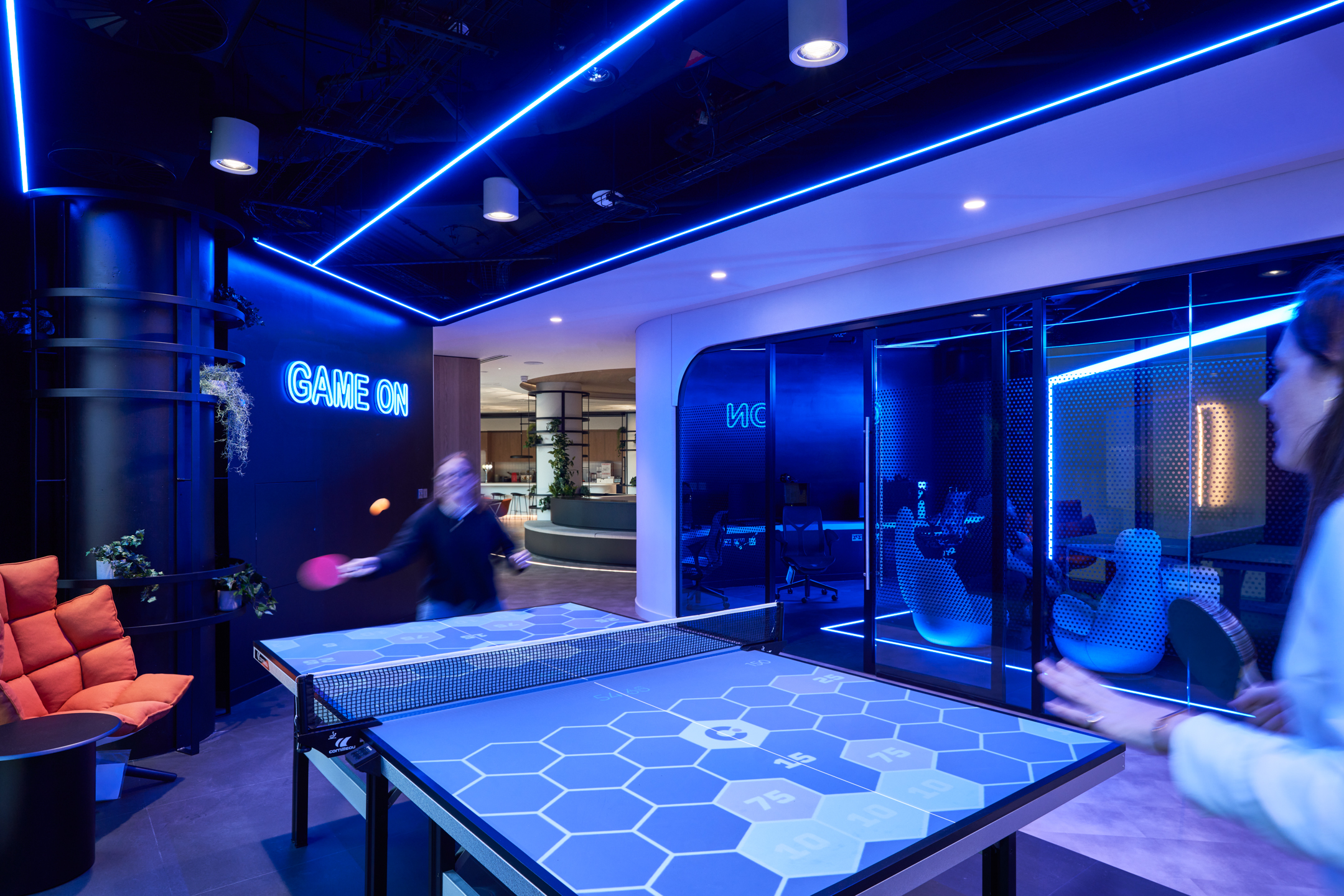 Capital.com office games room designed by Modus Workspace, featuring a futuristic, neon-lit space with a digital ping pong table and comfortable seating. The room is accented by bold blue lighting and a 'Game On' neon sign, creating an energetic and modern environment for employee relaxation and social interaction