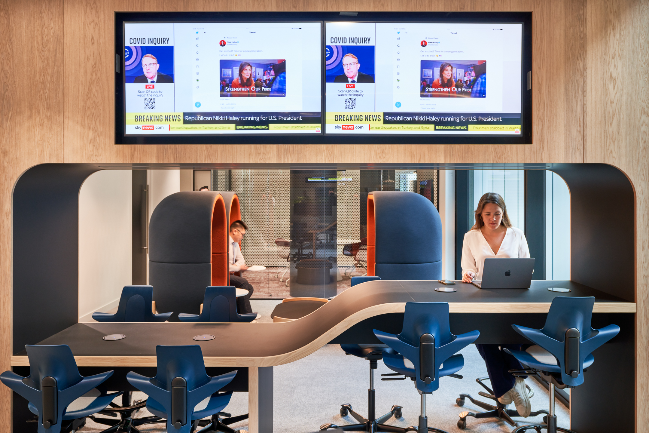 Capital.com office designed by Modus Workspace, featuring a modern work area with open desks, ergonomic chairs, and greenery. Two people are engaged in a conversation in built-in booths with bold, textured orange wall panels, while another individual works in a private meeting booth. The space is designed to promote collaboration and focus.