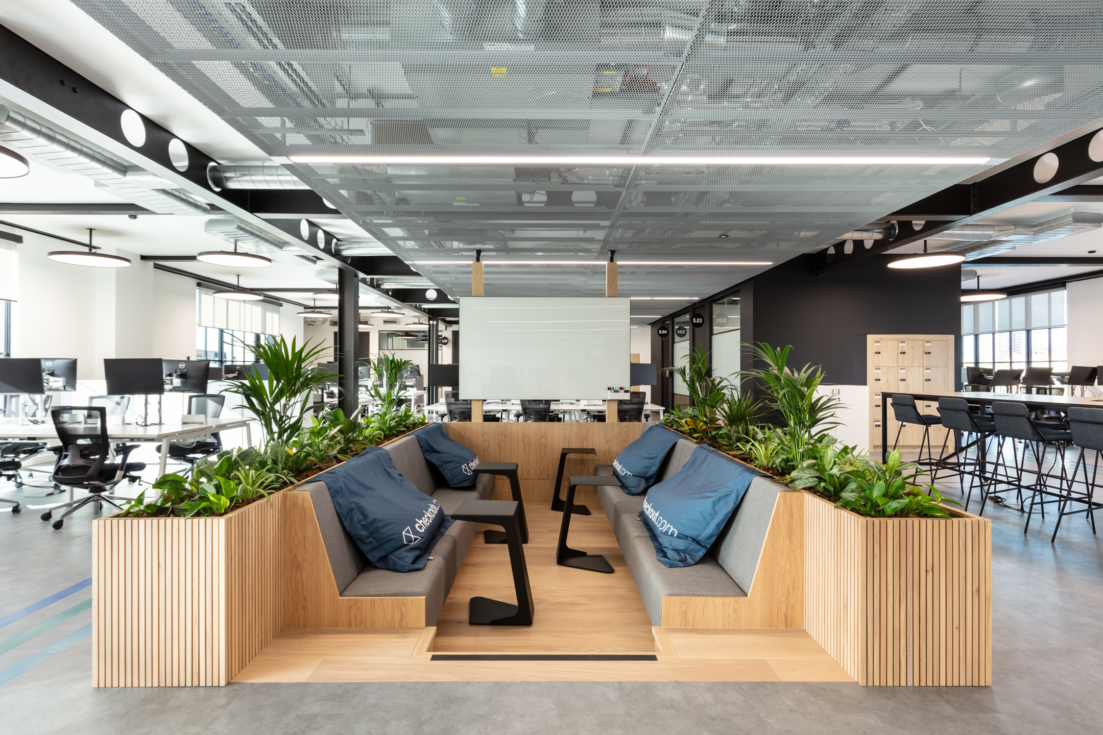 Checkout.com’s open-plan workspace, shortlisted in the SBID Awards 2021. The space has a central collaborative hub with benches and stools, surrounded by wood planters and green plants.