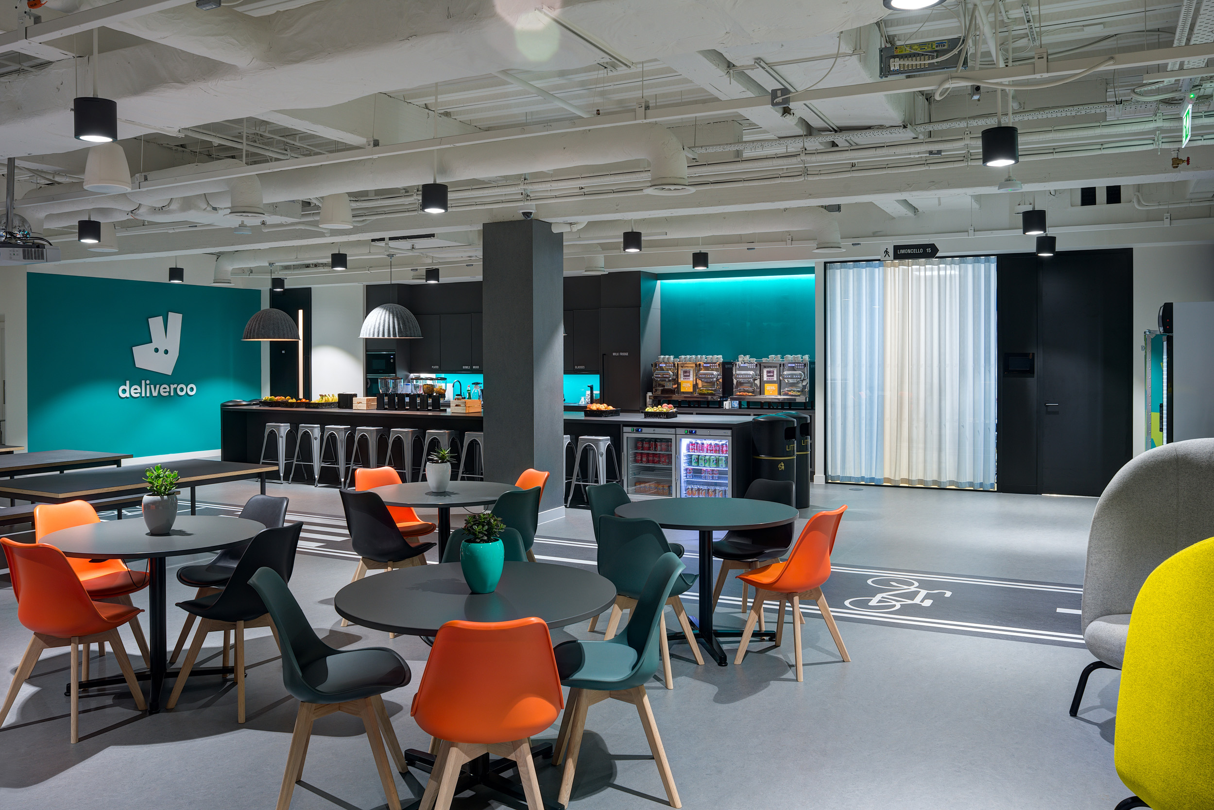 The teapoint at Deliveroo's London office with modern lighting, bar seating, and a vibrant teal backdrop. This area is designed for collaboration and informal meetings.