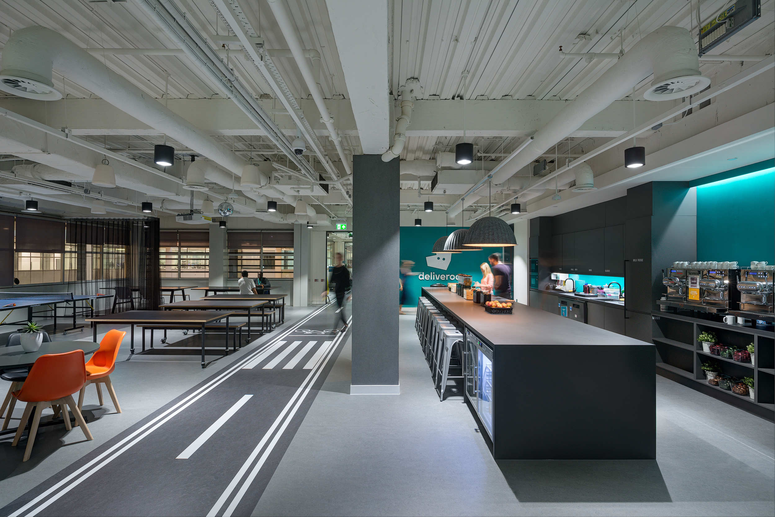 A view of the Deliveroo office featuring a spacious teapoint with bar seating and a vibrant workspace. The area includes a ping pong table and ample seating.