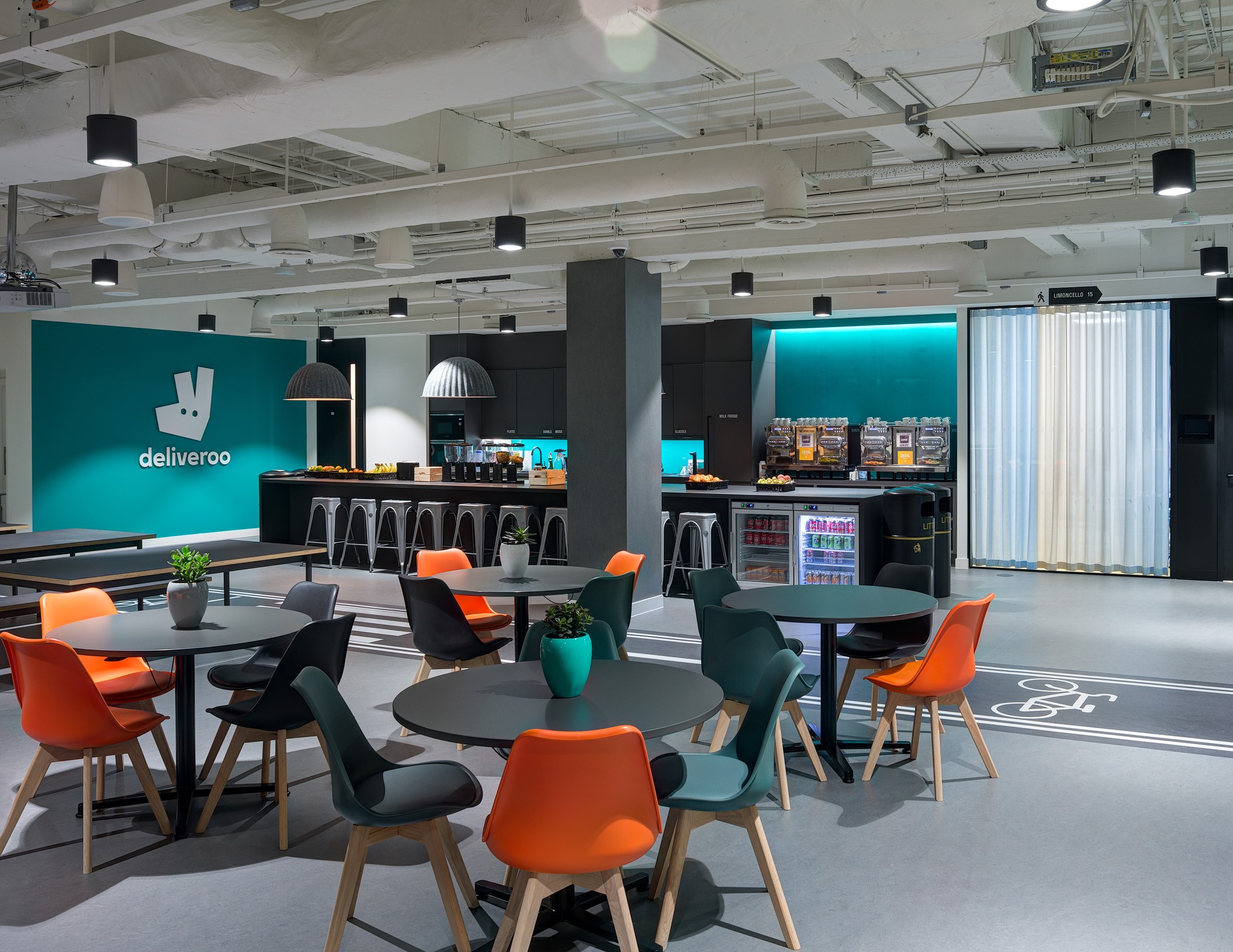 The teapoint at Deliveroo's London office with modern lighting, bar seating, and a vibrant teal backdrop. This area is designed for collaboration and informal meetings.