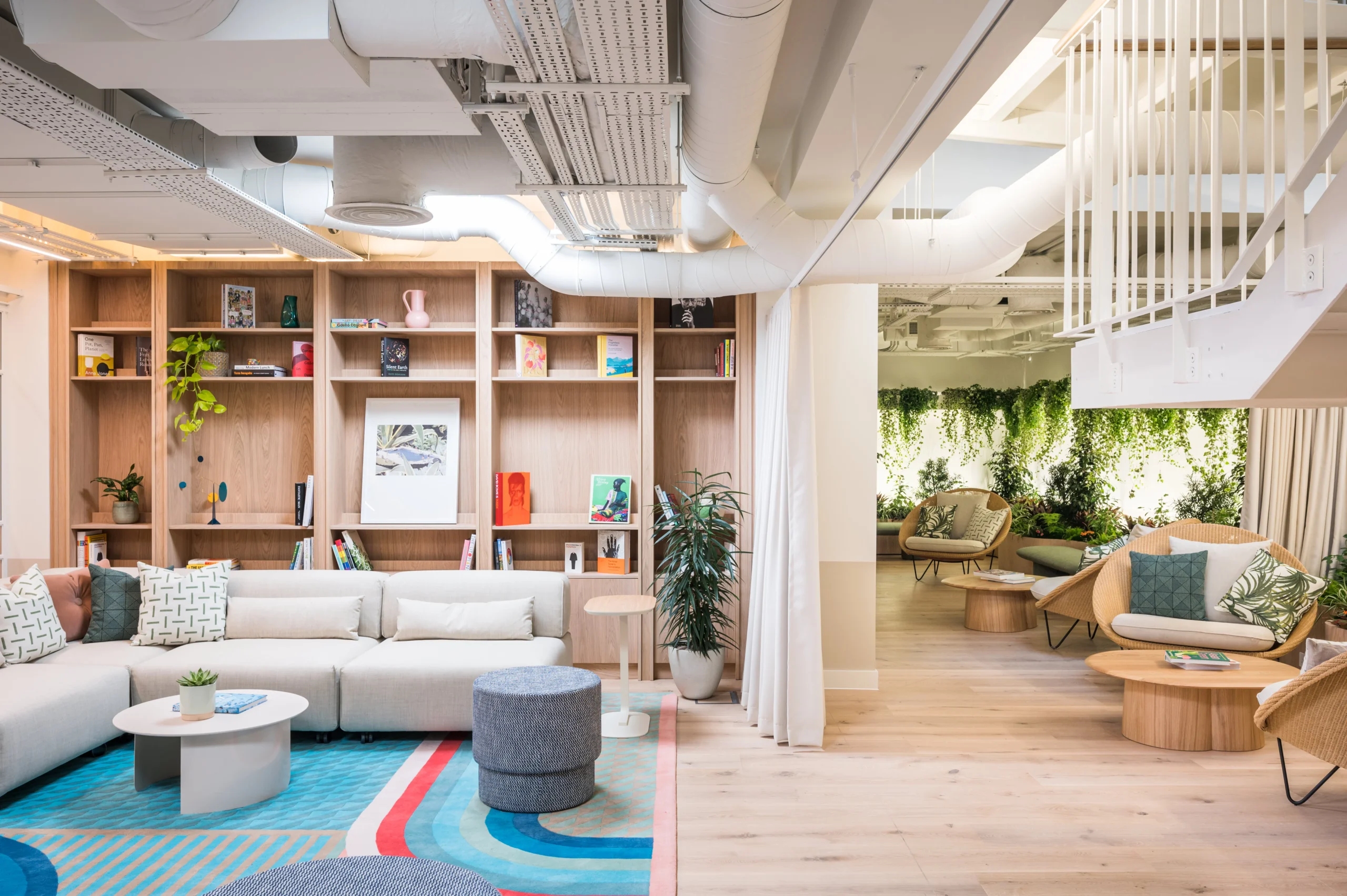 A contemporary, open office lounge area designed by Modus for Fora, featuring a colourful, patterned rug with cosy seating arrangements, including a sectional sofa and round poufs. Adjacent to the lounge is a staircase leading to a serene space filled with natural light and greenery, offering additional seating in a relaxed, biophilic design setting.