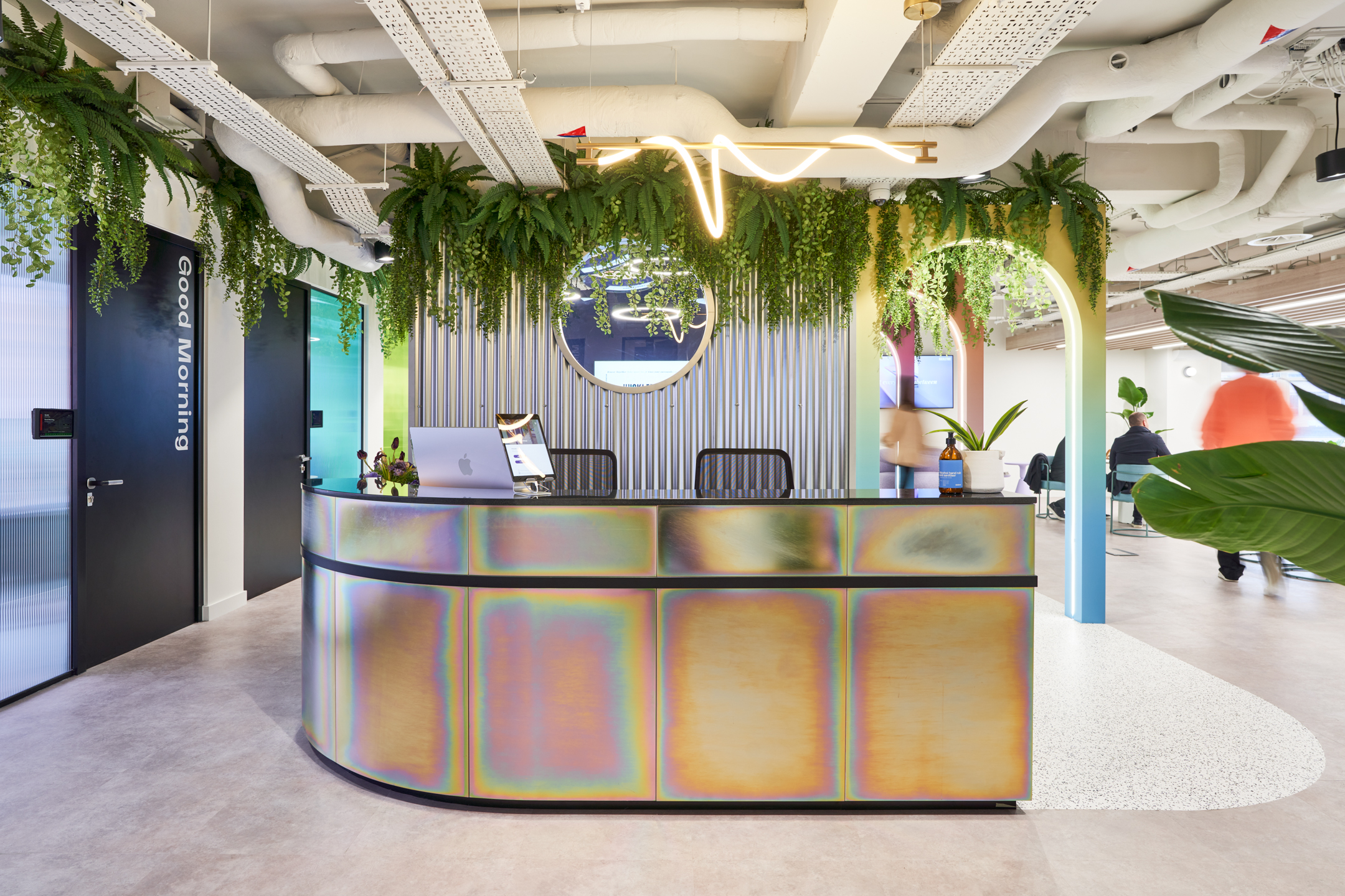 The reception area at Huckletree, featuring a sleek iridescent desk, surrounded by hanging greenery and illuminated with modern lighting, creating a welcoming and stylish entrance.