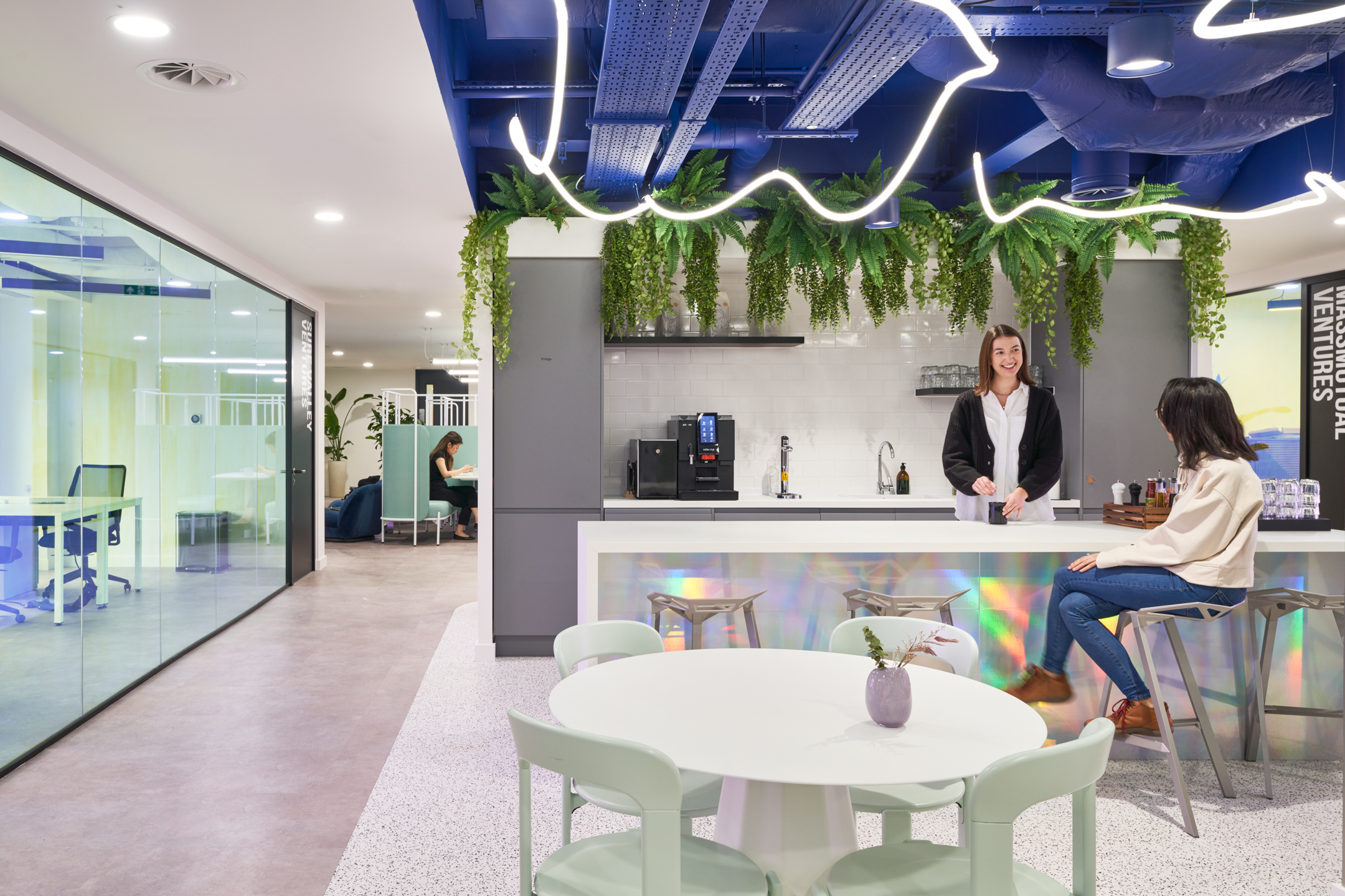 A modern kitchen and breakout area at Huckletree, featuring hanging greenery, sleek bar seating, and pastel-toned furniture, creating a relaxed and inviting space for socialising.