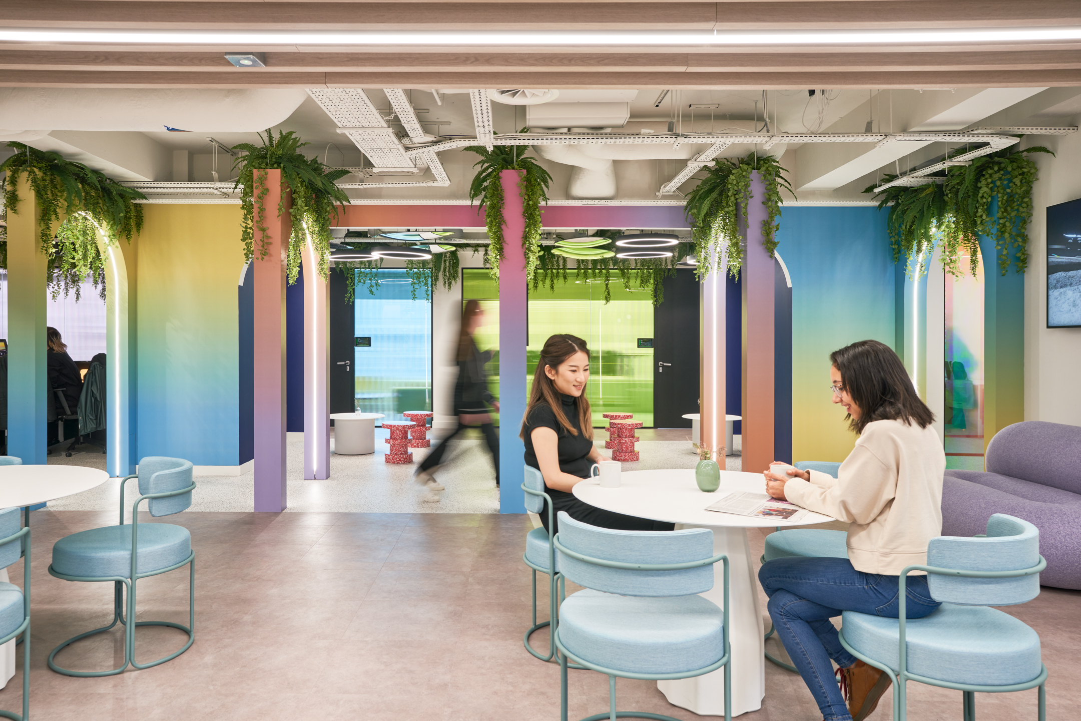 A vibrant communal area at Huckletree, featuring pastel-coloured seating, white tables, and rainbow-toned archways adorned with hanging greenery, creating a lively workspace.