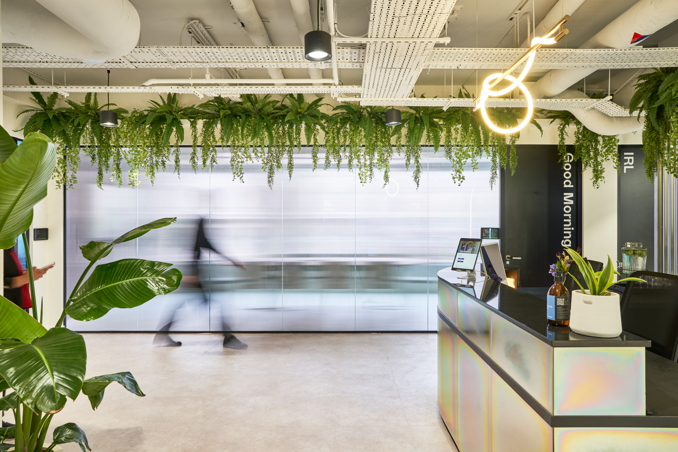 Huckletree’s sleek reception area with an iridescent desk, hanging greenery from the ceiling, and modern lighting fixtures, creating a dynamic and welcoming atmosphere.