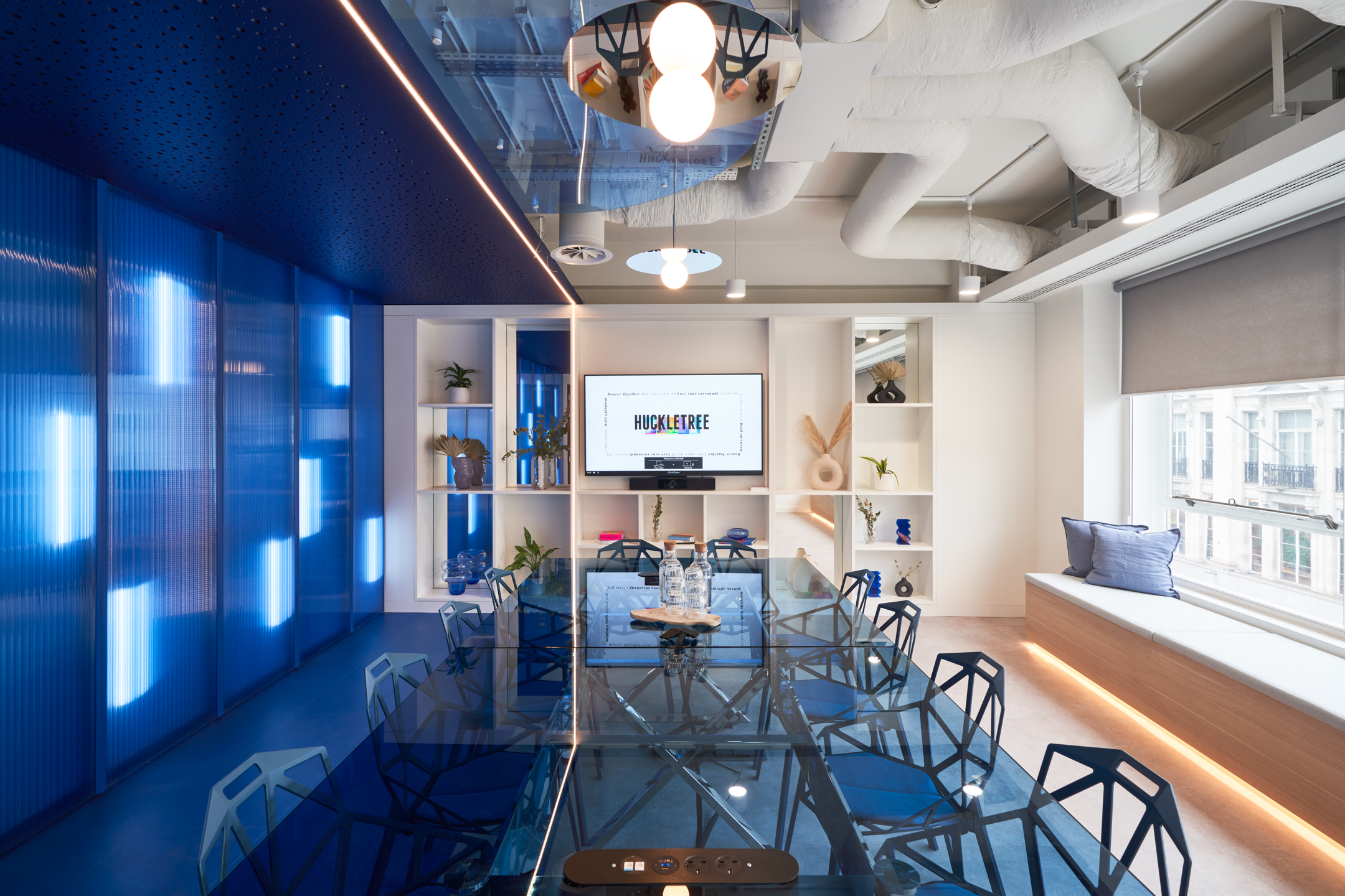 A stylish meeting room at Huckletree, featuring a sleek glass table, geometric chairs, blue accent lighting, and a display screen, creating a modern and professional setting.