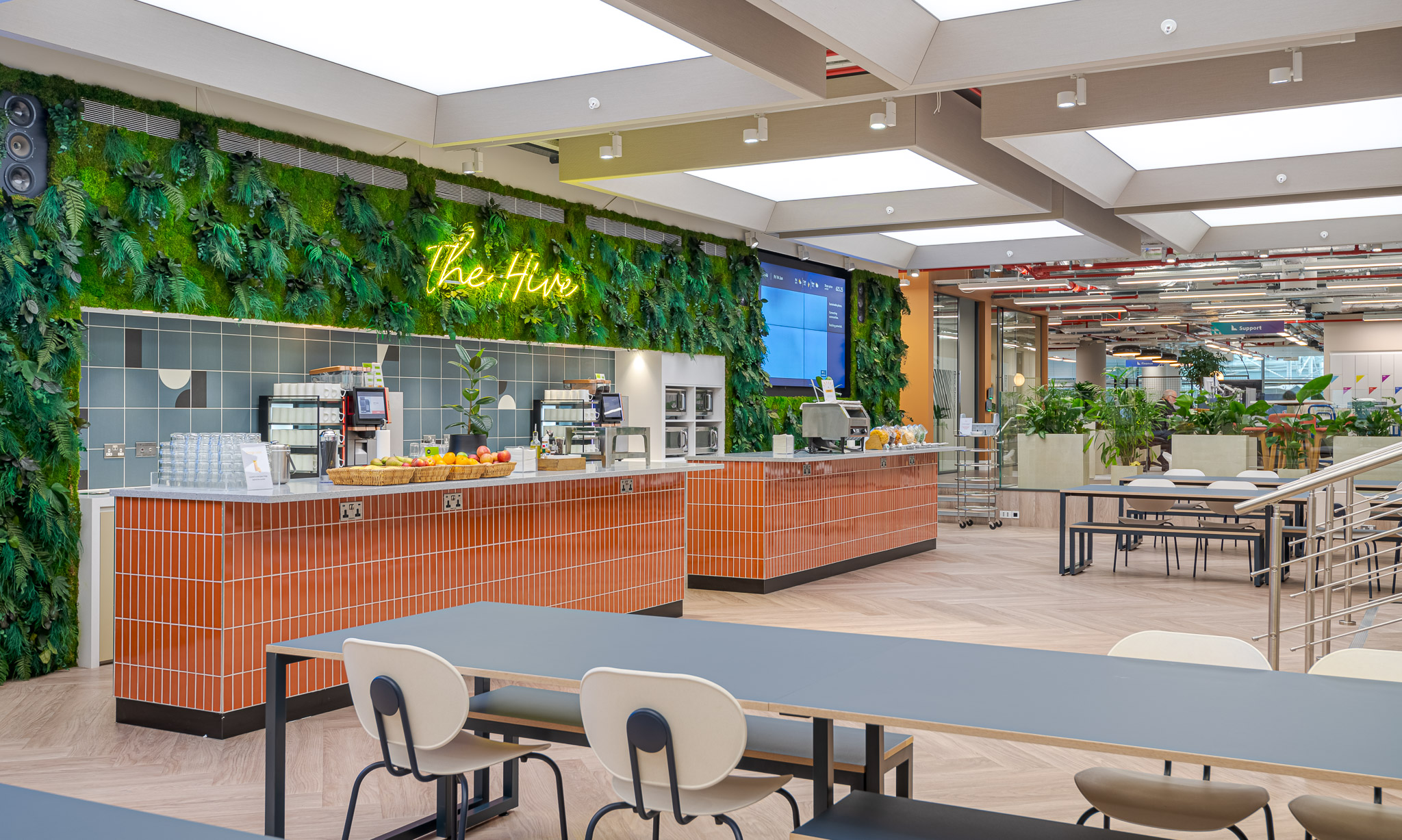 Bright and stylish communal dining area in Landsec's 100 Victoria Street Headquarters, designed by Modus Workspace. The area showcases a green living wall, sleek, orange-tiled counters, and ample seating for employees.