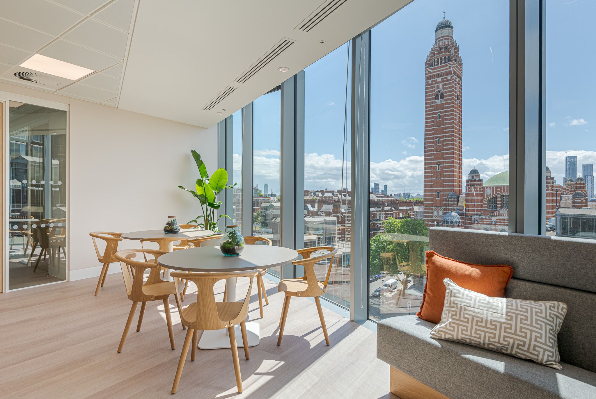 Tenant-ready lounge at Landsec 100 Victoria Level 6 designed by Modus Workspace. Featuring round tables and wooden chairs with panoramic views of London landmarks through floor-to-ceiling windows, perfect for casual meetings or breaks.