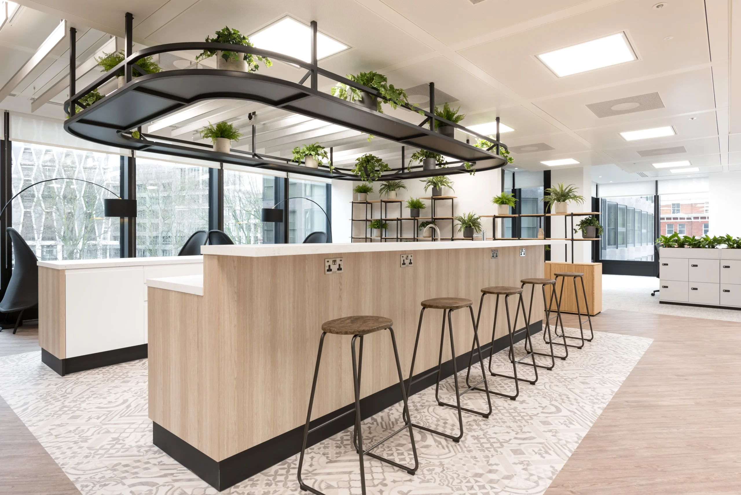 A modern office space at London and Oriental designed by Modus Workspace. The space includes a sleek kitchen area with a wooden island, black bar stools, and an overhead structure adorned with greenery, emphasizing a biophilic design approach.