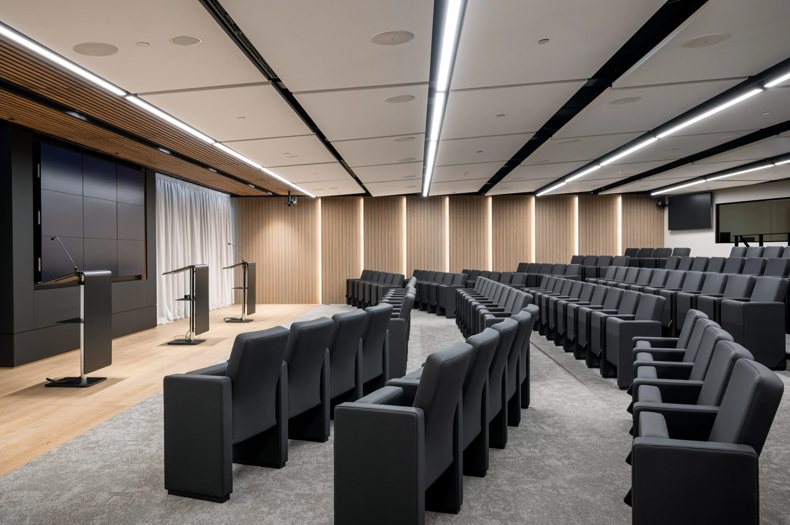 Numis’ modern well-lit auditorium with tiered seating, featuring rows of cushioned dark grey chairs. At the front are podiums and a large screen with wood panelling and warm white LED lights on the wall.