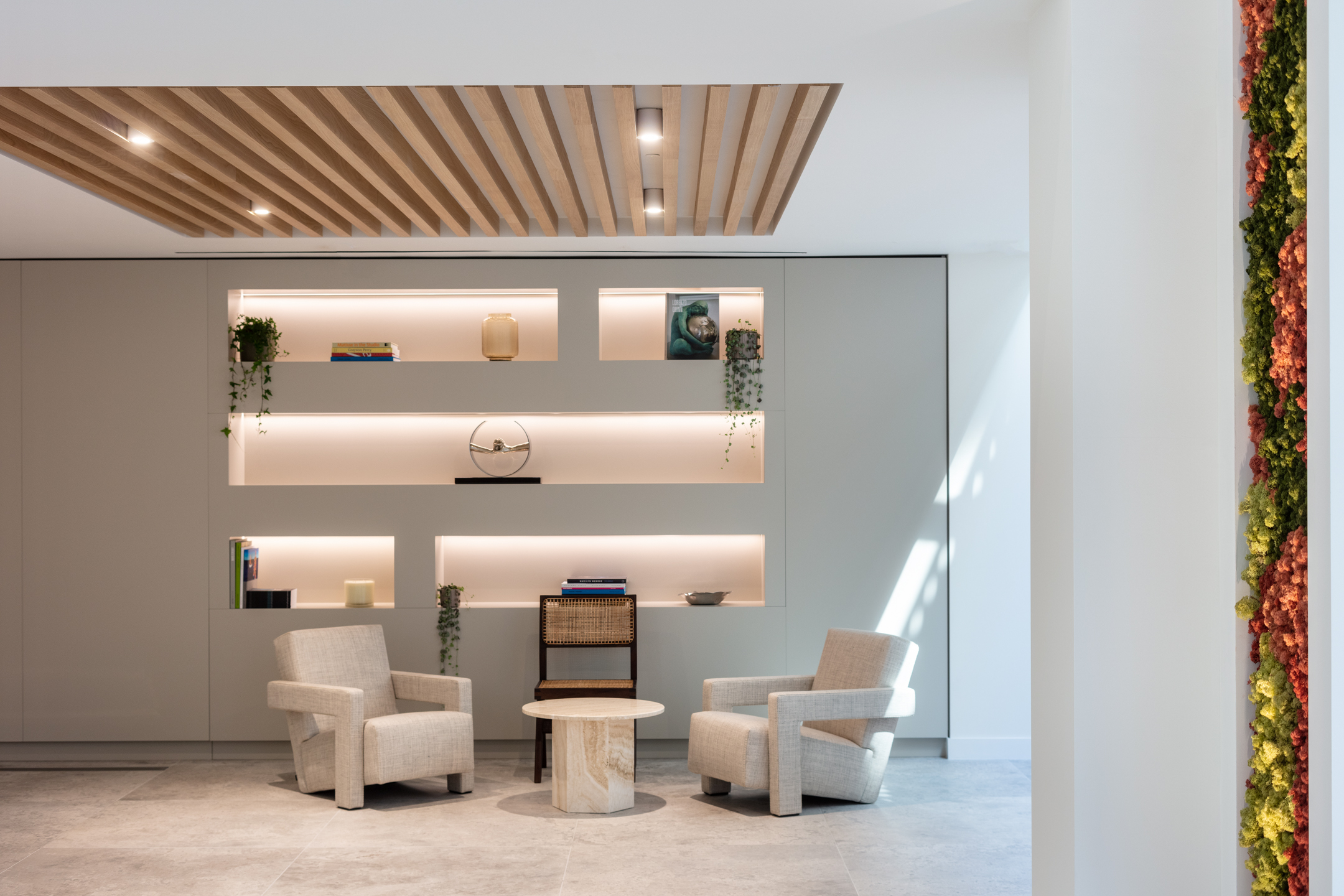 A serene seating area in the Ouronyx aesthetic clinic in Mayfair, designed by Modus Workspace, featuring two plush armchairs and a small marble table. The space is accented by minimalist, backlit shelving and a slatted wood ceiling detail, creating a calm and inviting environment