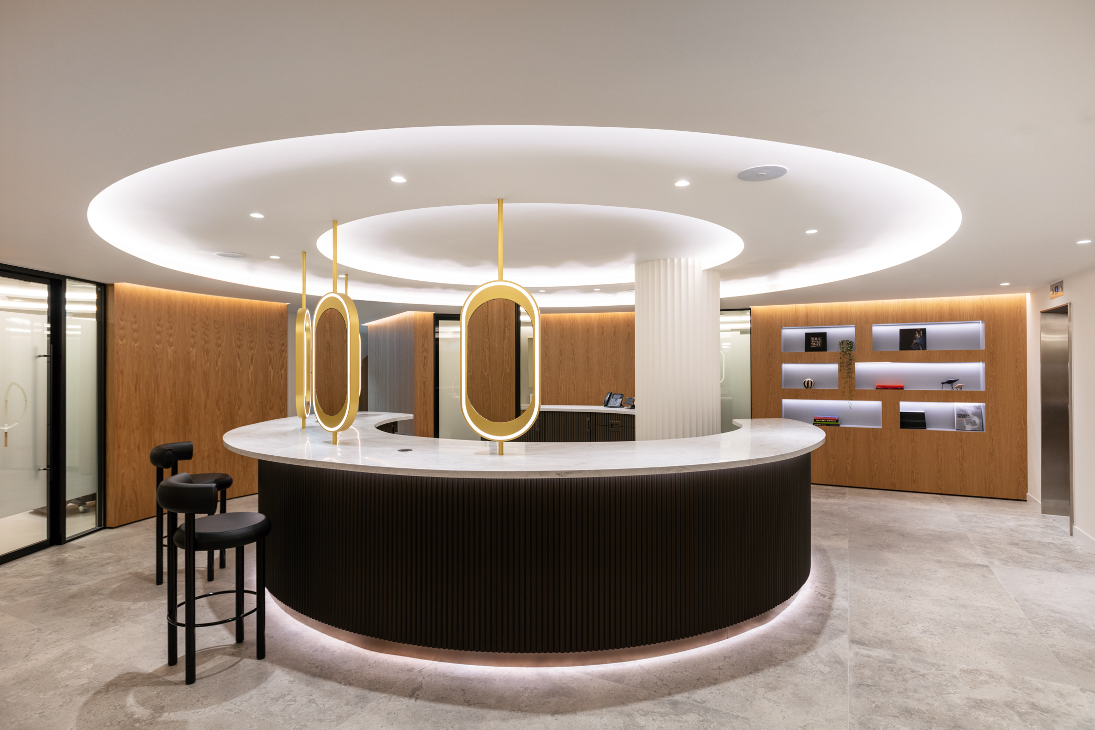 A sophisticated curved beauty bar in the Ouronyx aesthetic clinic in Mayfair, designed by Modus Workspace, featuring a curved marble counter, backlit by soft ambient lighting. The space includes elegant circular mirrors suspended from the ceiling, wood paneling, and sleek shelving, creating a high-end, contemporary atmosphere.