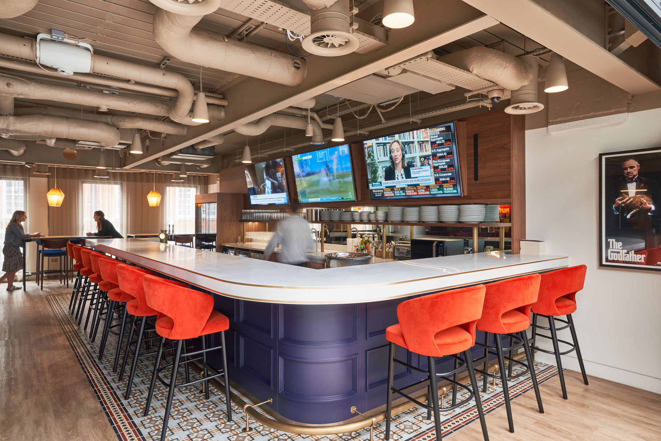 A sophisticated bar area at a private equity firm in Marylebone, designed by Modus Workspace. Bold red barstools surround a sleek marble countertop, with people engaging in conversations in the background, enhancing the workspace’s social atmosphere.