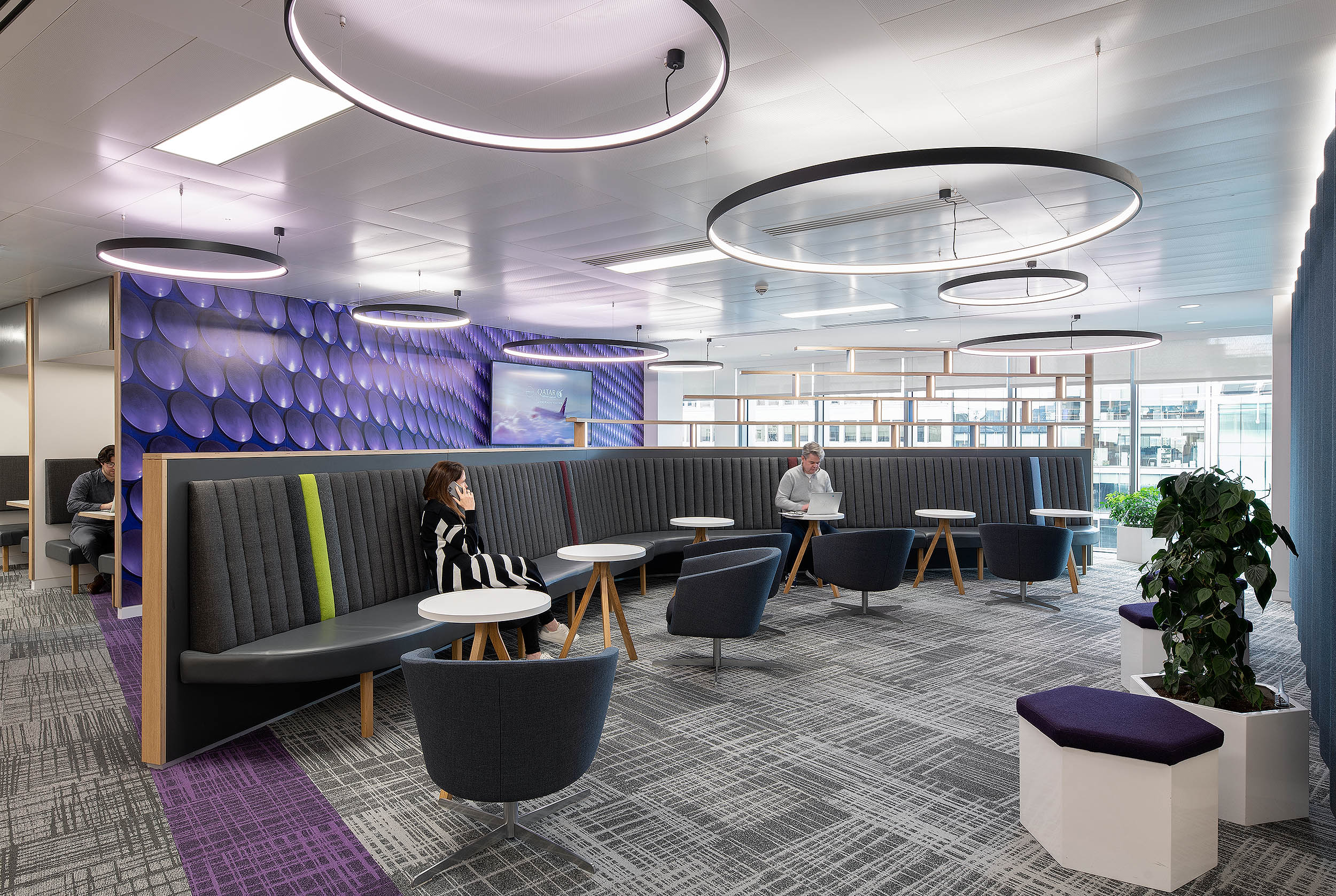 Breakout area at RICS with booth-style seating, round tables, and a feature wall with purple textures, set under modern lighting fixtures.