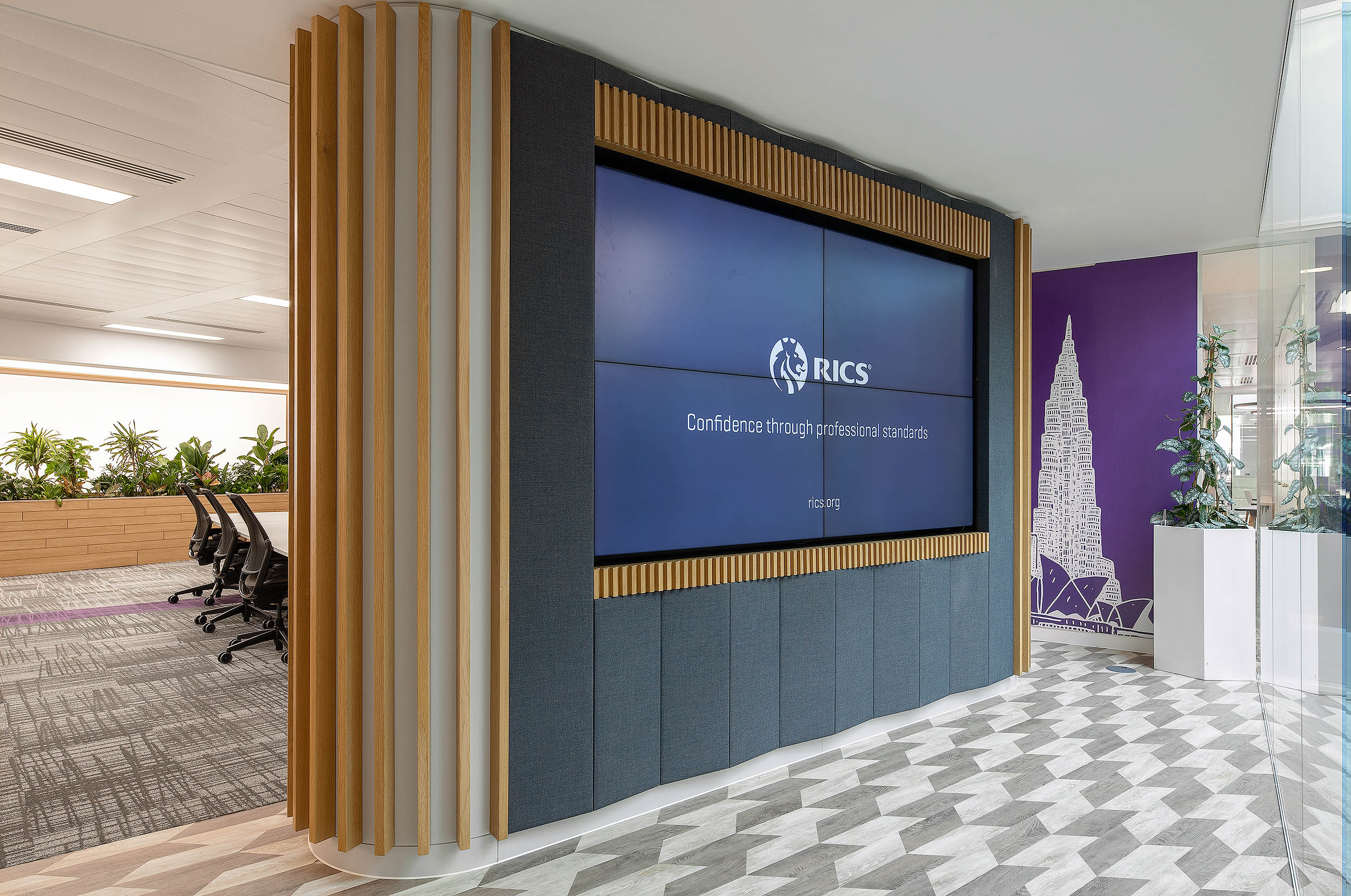 The large digital screen at RICS reception is framed by vertical wooden slats adjacent to a boardroom with grey chairs and a plant feature