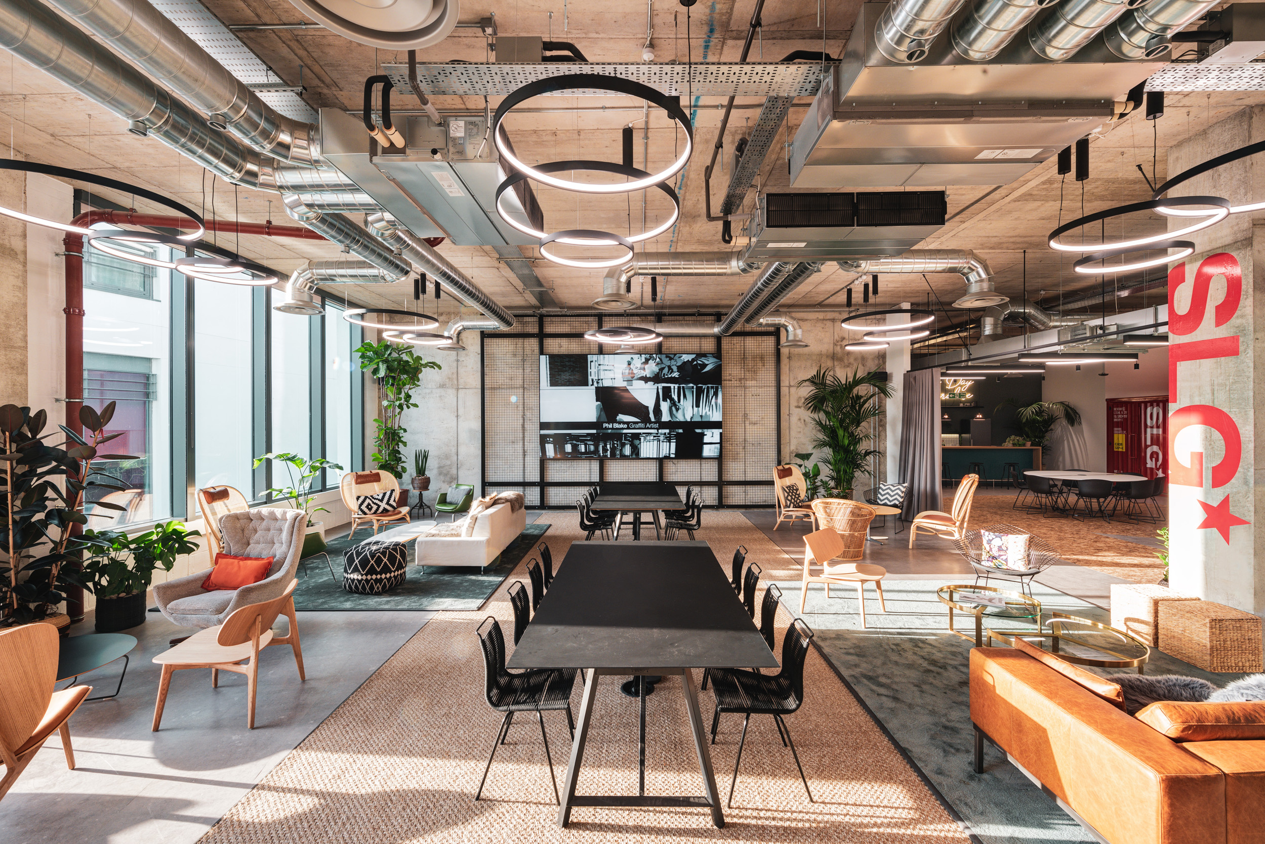 Industrial-inspired collaborative workspace at SLG designed by Modus, featuring exposed ducts, modern pendant lighting, and versatile seating areas. The open-plan layout incorporates greenery and warm tones, blending functionality with comfort for an innovative work environment.