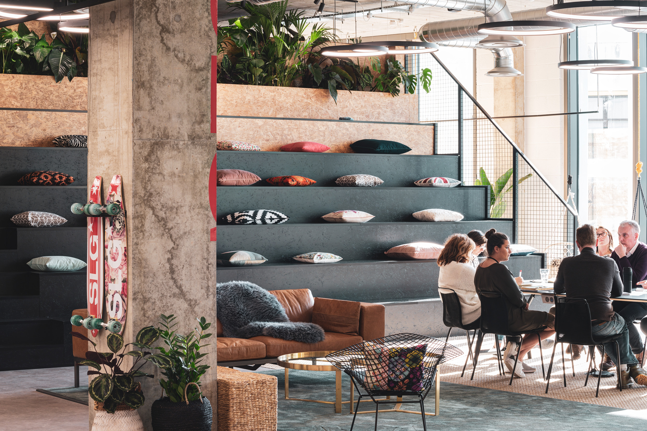 Open, relaxed workspace featuring tiered seating with colourful cushions, a casual meeting area, and indoor plants, designed to transition into a flexible events space where necessary.