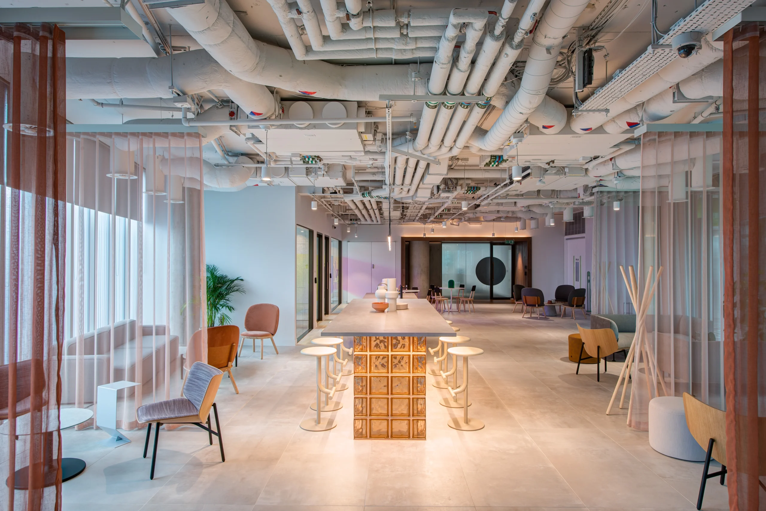 Spacious open lounge area at Storey - 2 Kingdom Street, designed by Modus Workspace. The space features high ceilings, exposed pipework, and comfortable seating, creating a welcoming environment for collaboration and relaxation. A long table with modern stools acts as a central gathering point.