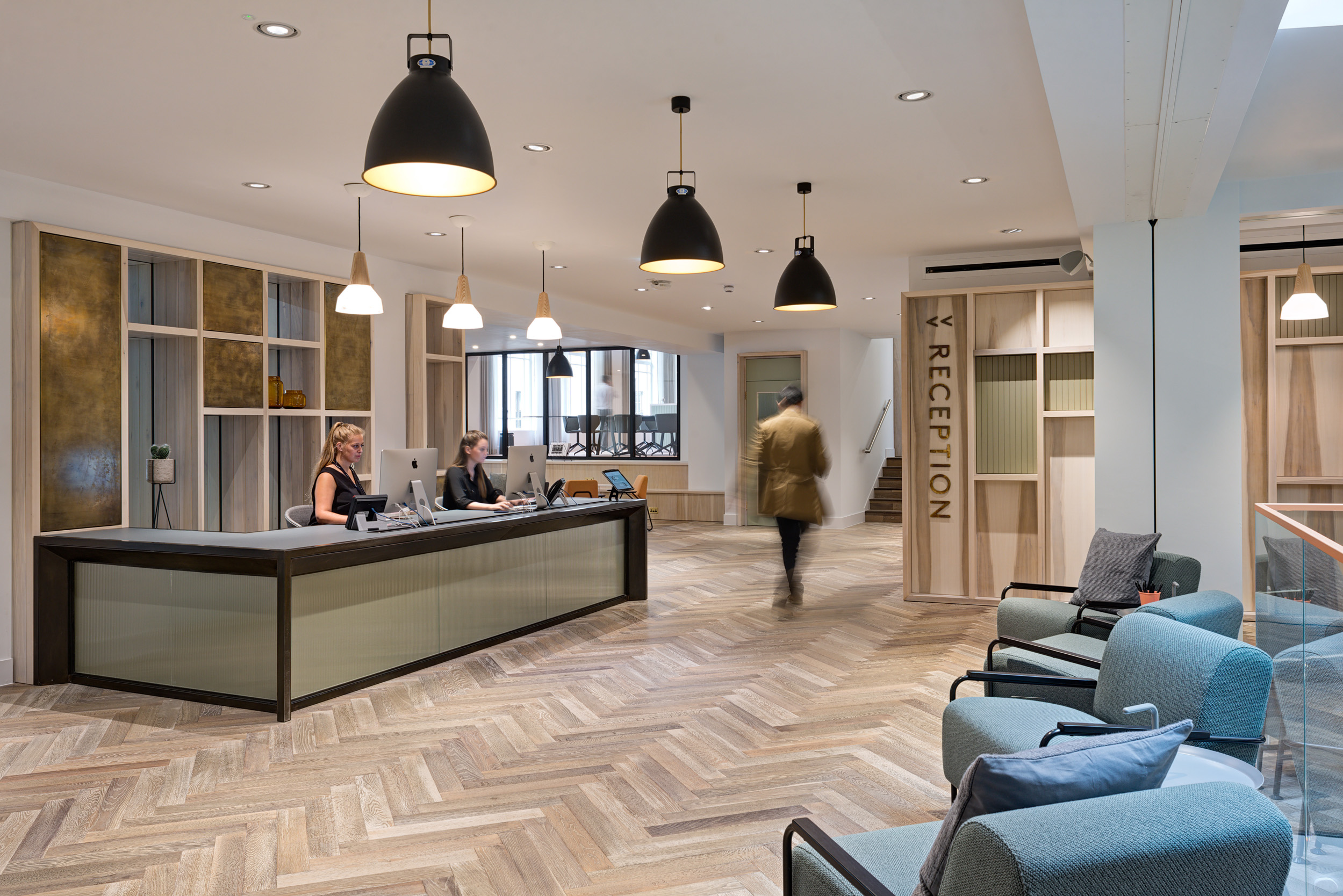 A modern reception area at The Office Group Wimpole Street, designed by Modus Workspace. The sleek desk is attended by staff while another person approaches. The room features pendant lights and comfortable seating in soft, muted tones.