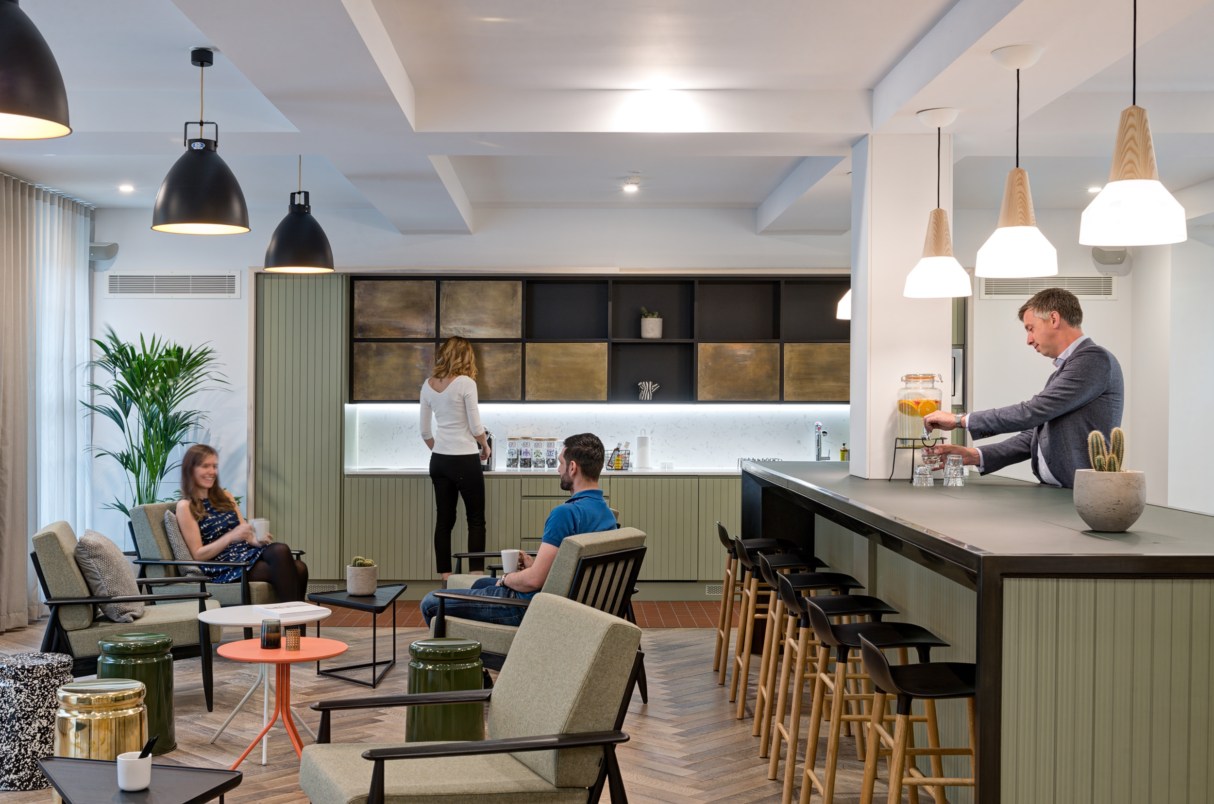 A cosy teapoint with seating at The Office Group Wimpole Street, designed by Modus Workspace. People are enjoying coffee in the lounge, highlighted by stylish lighting and earthy green tones