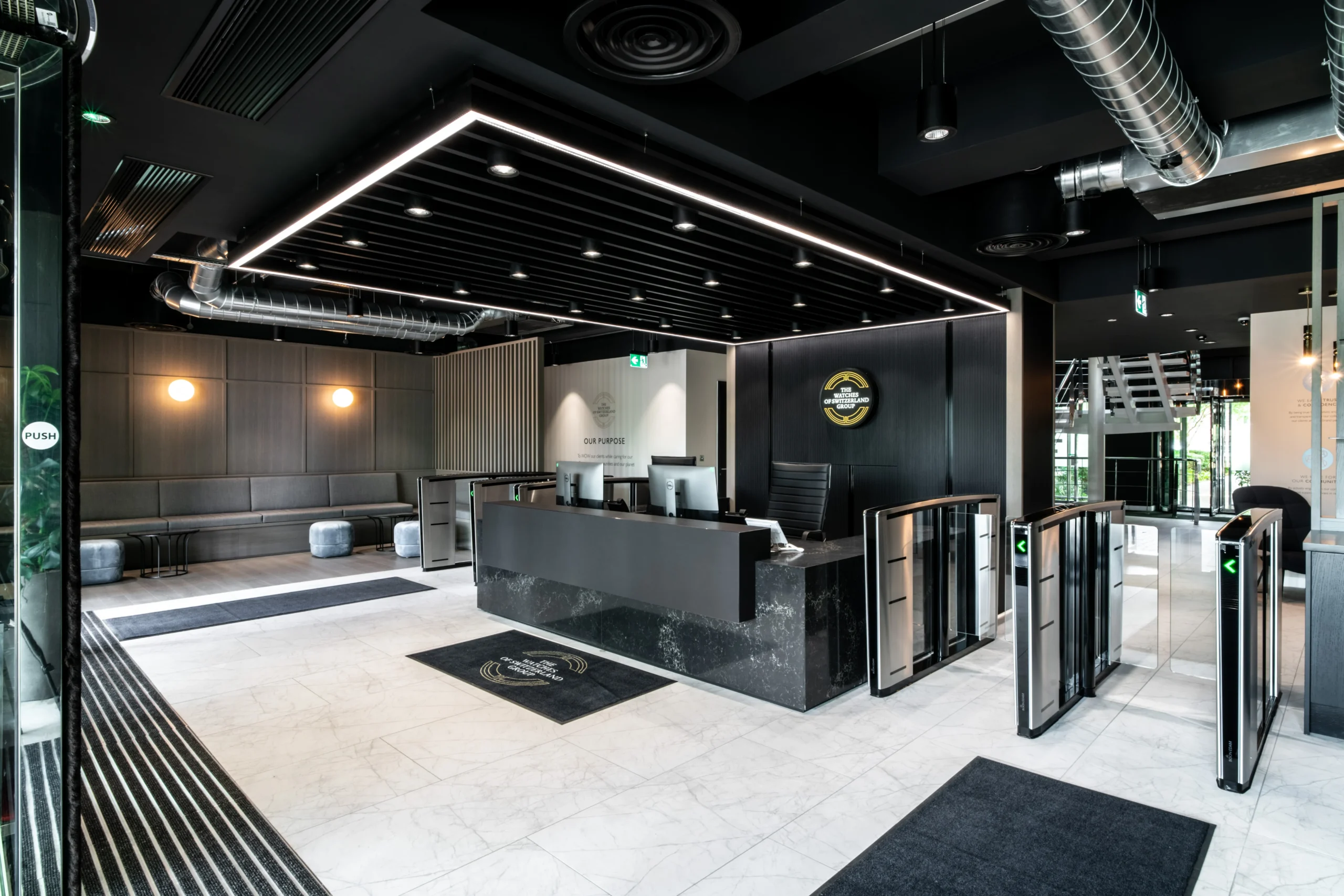 Modern reception area with sleek black and grey tones, featuring a central desk and turnstile entrance for Watches of Switzerland Group, designed by Modus Workspace.