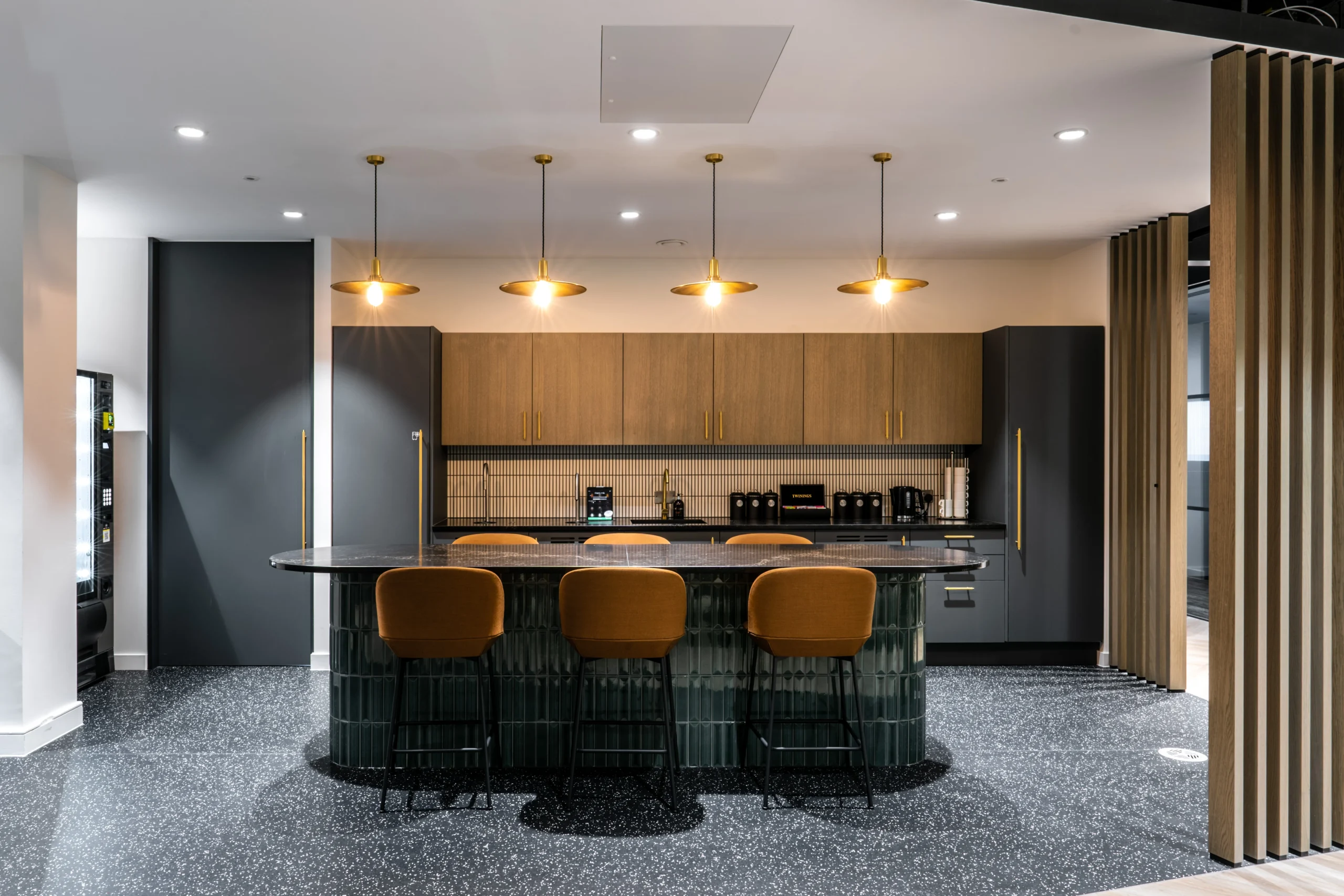 A contemporary teapoint area at the Watches of Switzerland Group headquarters featuring a sleek island with bar seating, warm wood cabinetry, and elegant lighting that creates a welcoming space for employees to connect and recharge.