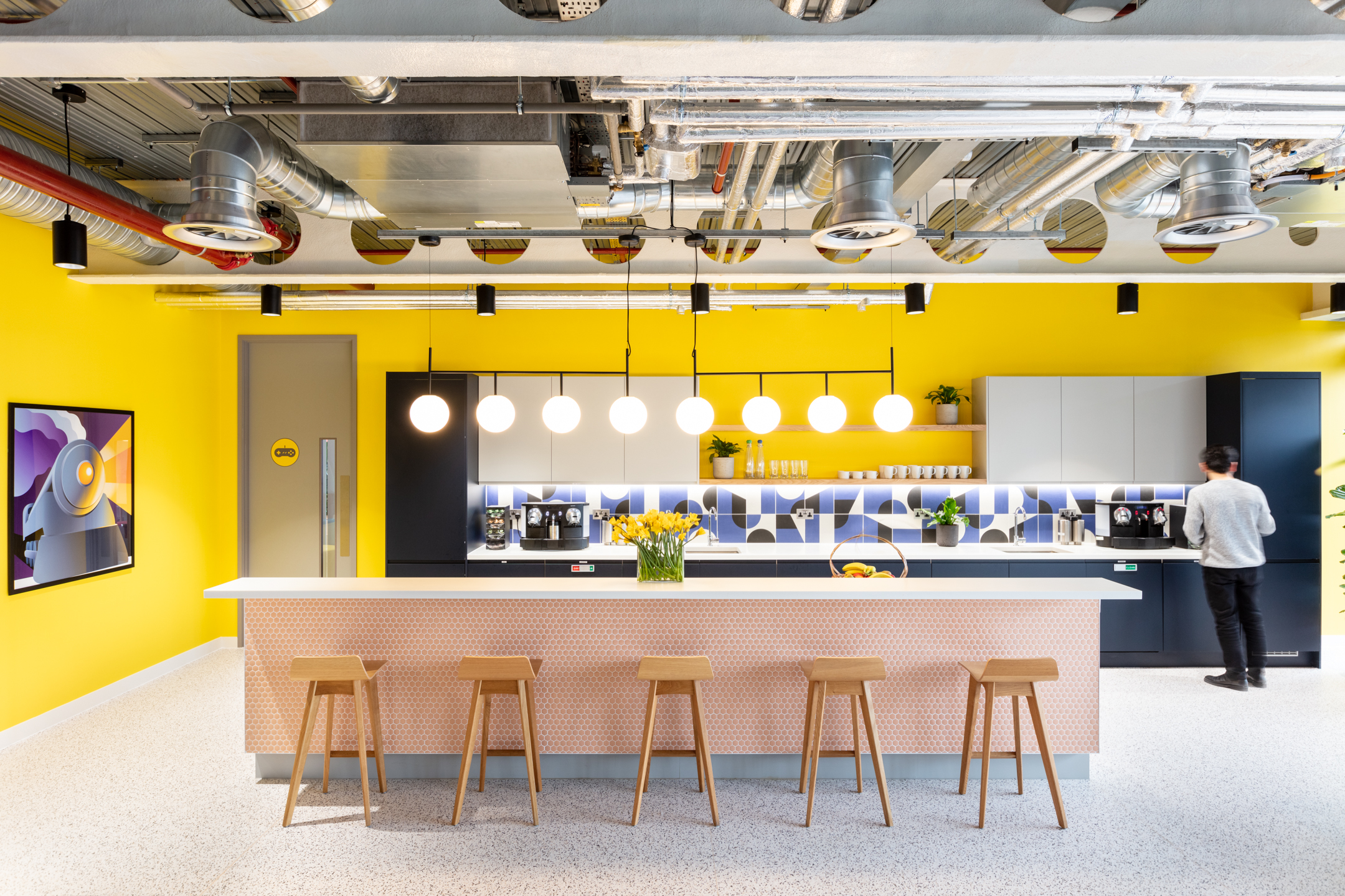 Travelfusion’s vibrant workspace Teapoint area with a bright yellow wall and a sleek, modern design. A long countertop with wooden stools serves as the central feature, beneath a row of hanging globe lights.