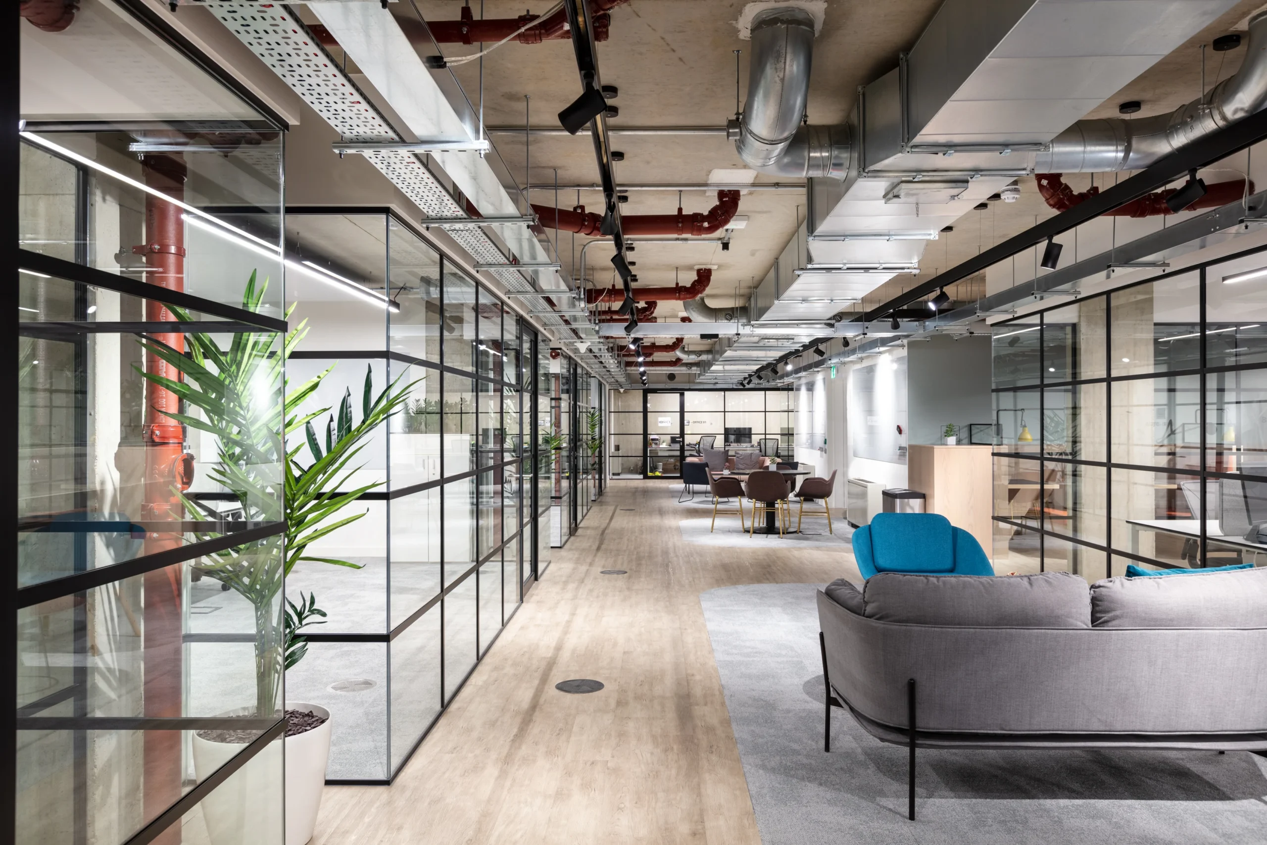 OSI’s spacious, open-plan workspace featuring glass-walled meeting rooms, exposed ceiling ducts, and modern furnishings, including a grey sofa and blue chair in a casual seating area.