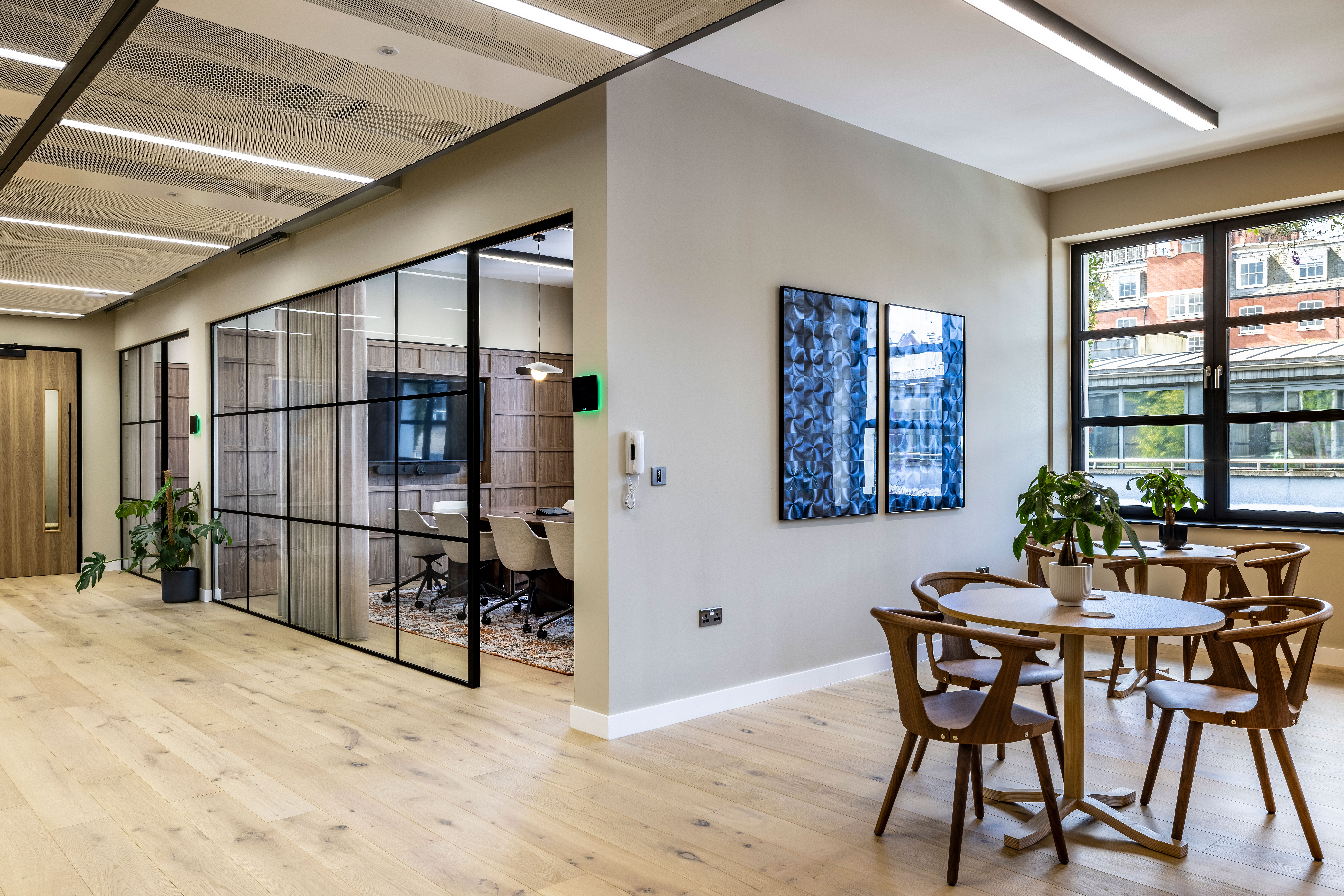 Transition’s London office showcasing a shared breakout area with wooden tables and chairs, and a glass-enclosed meeting room with natural light and modern decor.