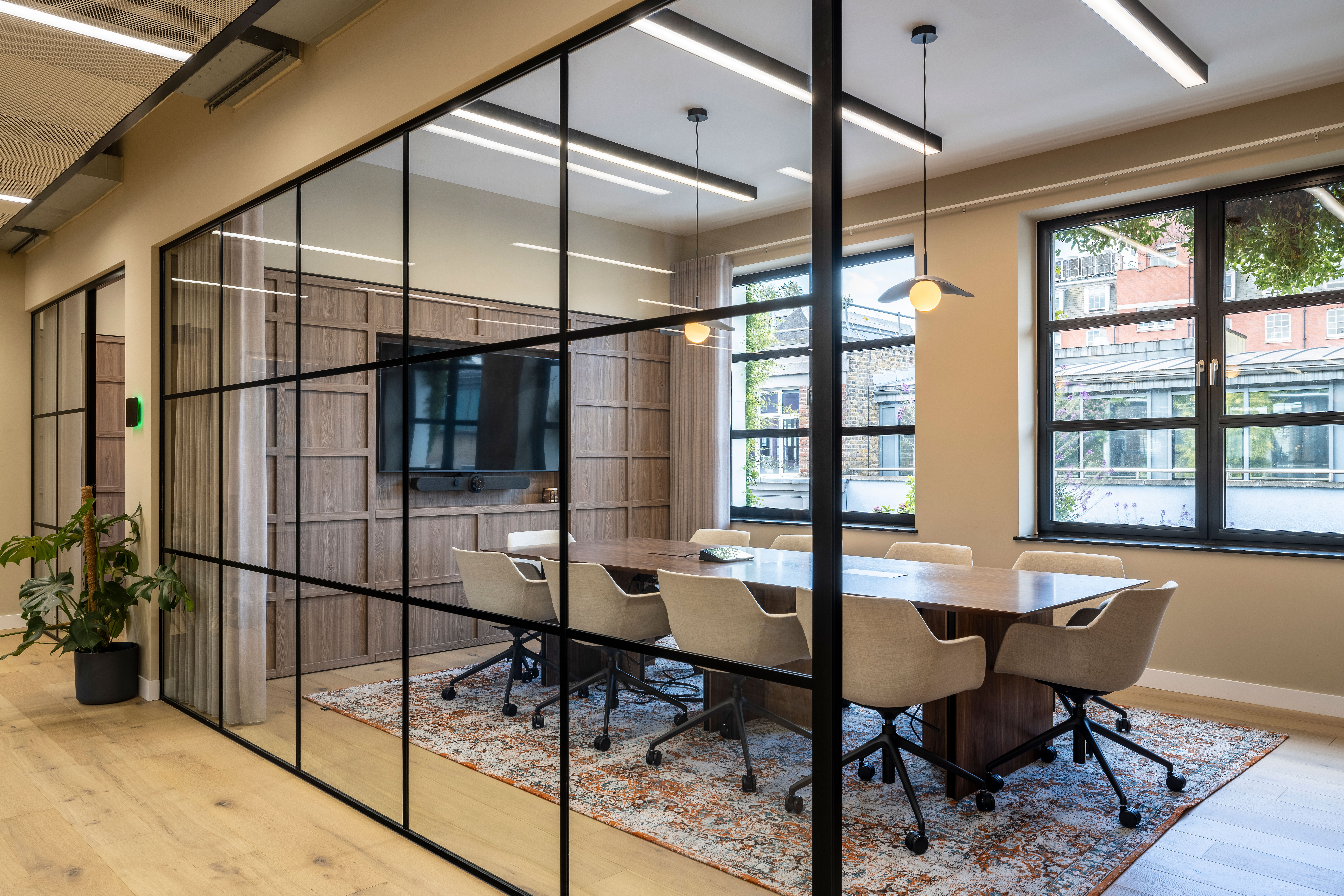 A meeting room in Transition’s London office, enclosed by glass walls, featuring a wooden table, upholstered chairs, a wall-mounted AV screen, and natural light.