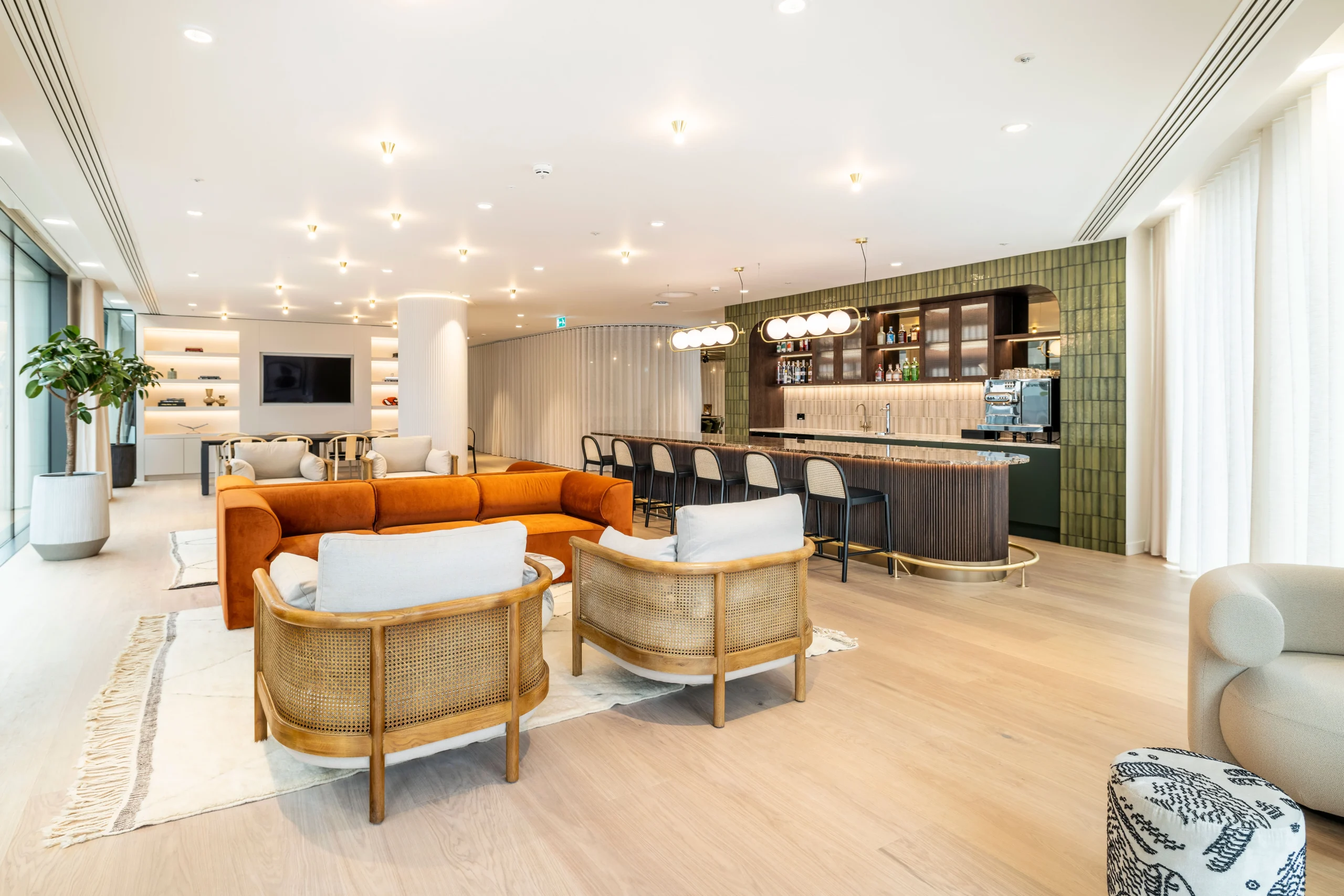 A modern bar area at Authentic Brand Group, designed by Modus, with a rich velvet orange sofa, marble tabletops, and a fully stocked bar accented by globe lighting.