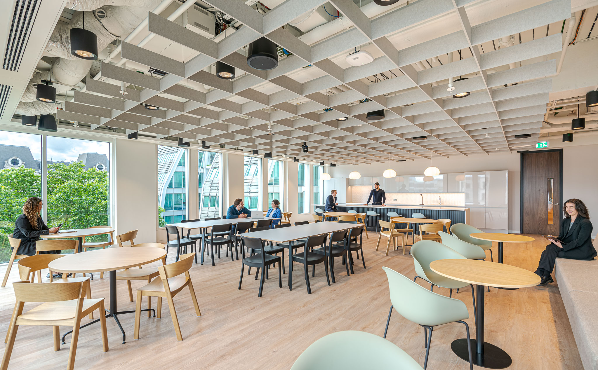 A modern open plan teapoint at the Bank of Ireland, designed by Modus Workspace, features minimalist wooden tables and chairs with large windows that offer views of the city skyline.