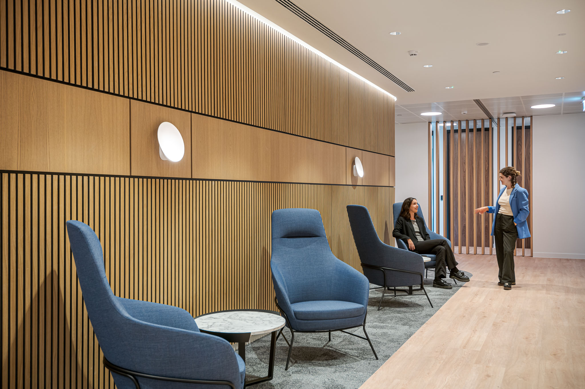 A quiet reception seating area at the Bank of Ireland, designed by Modus Workspace, features blue lounge chairs and wooden slatted walls, perfect for relaxed conversations.