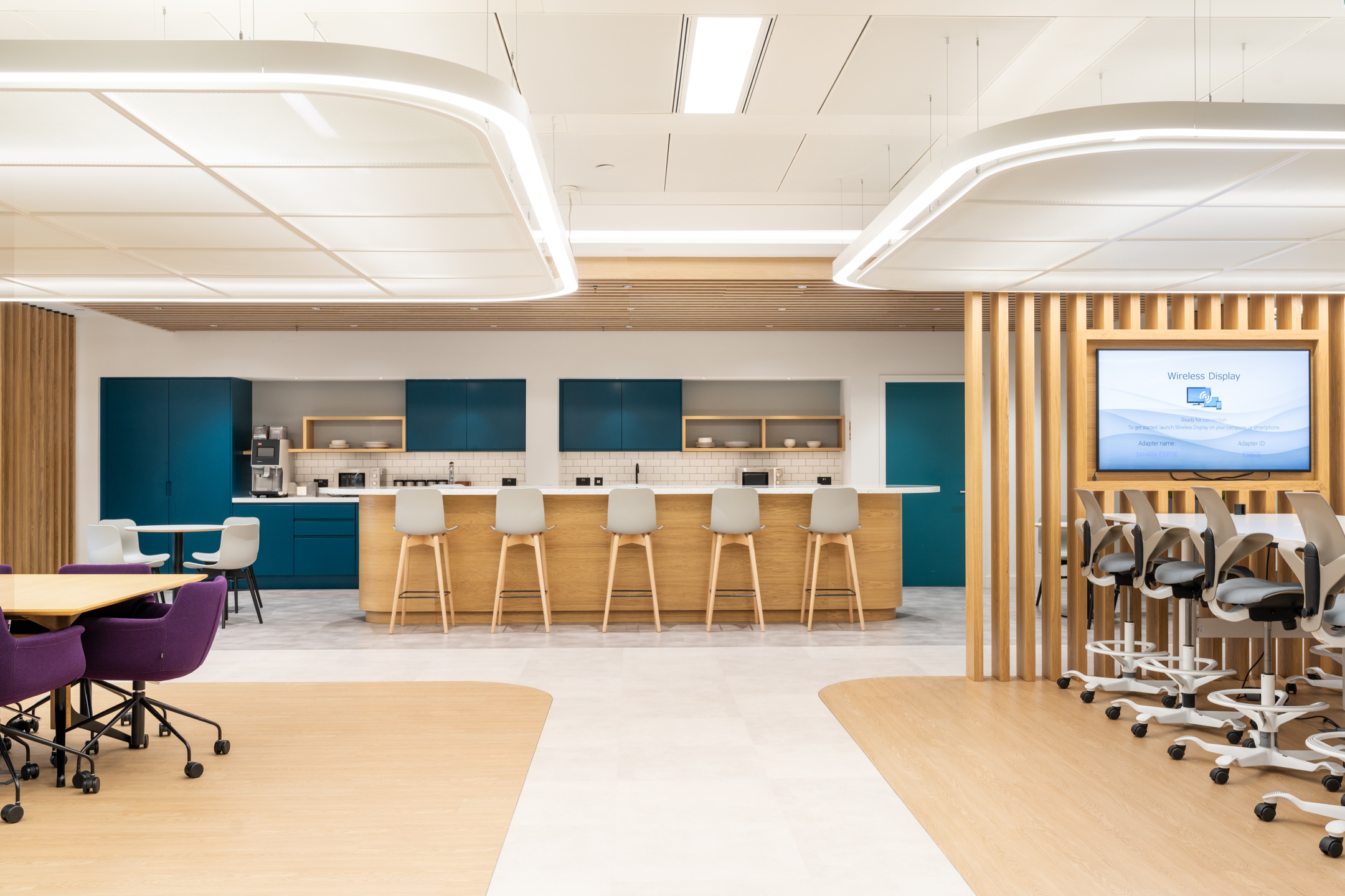 DXC Technology's modern teapoint features a long wooden island with high stools, teal cabinets, and bright lighting.