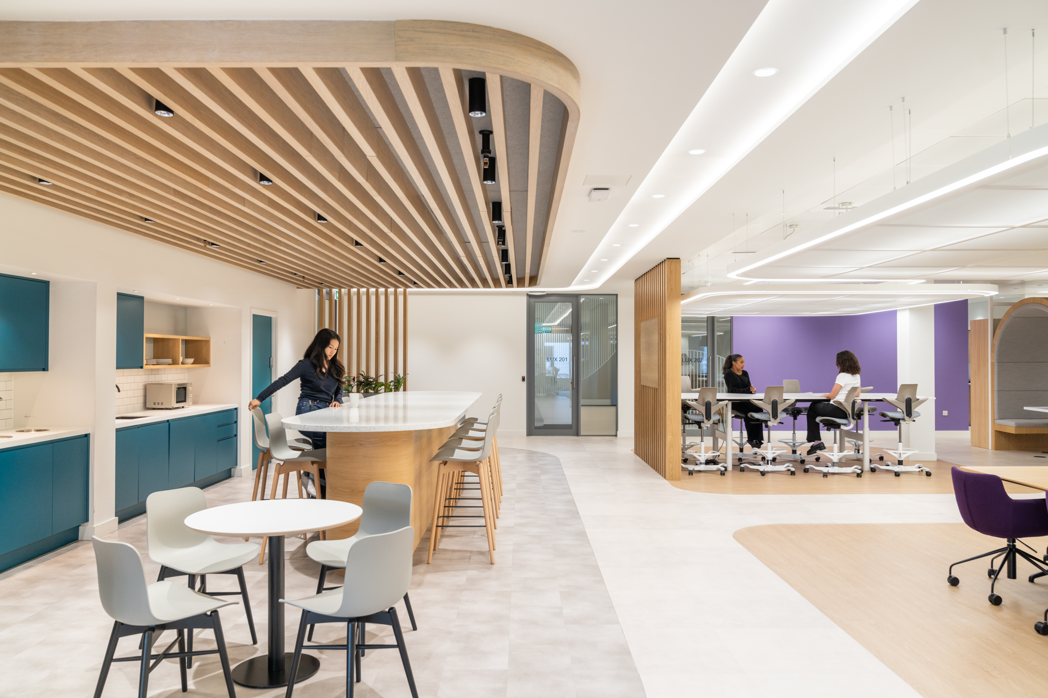 The collaborative workspace and teapoint at DXC Technology feature a long island, highchairs, and individual meeting areas with teal accents and warm wood finishes.