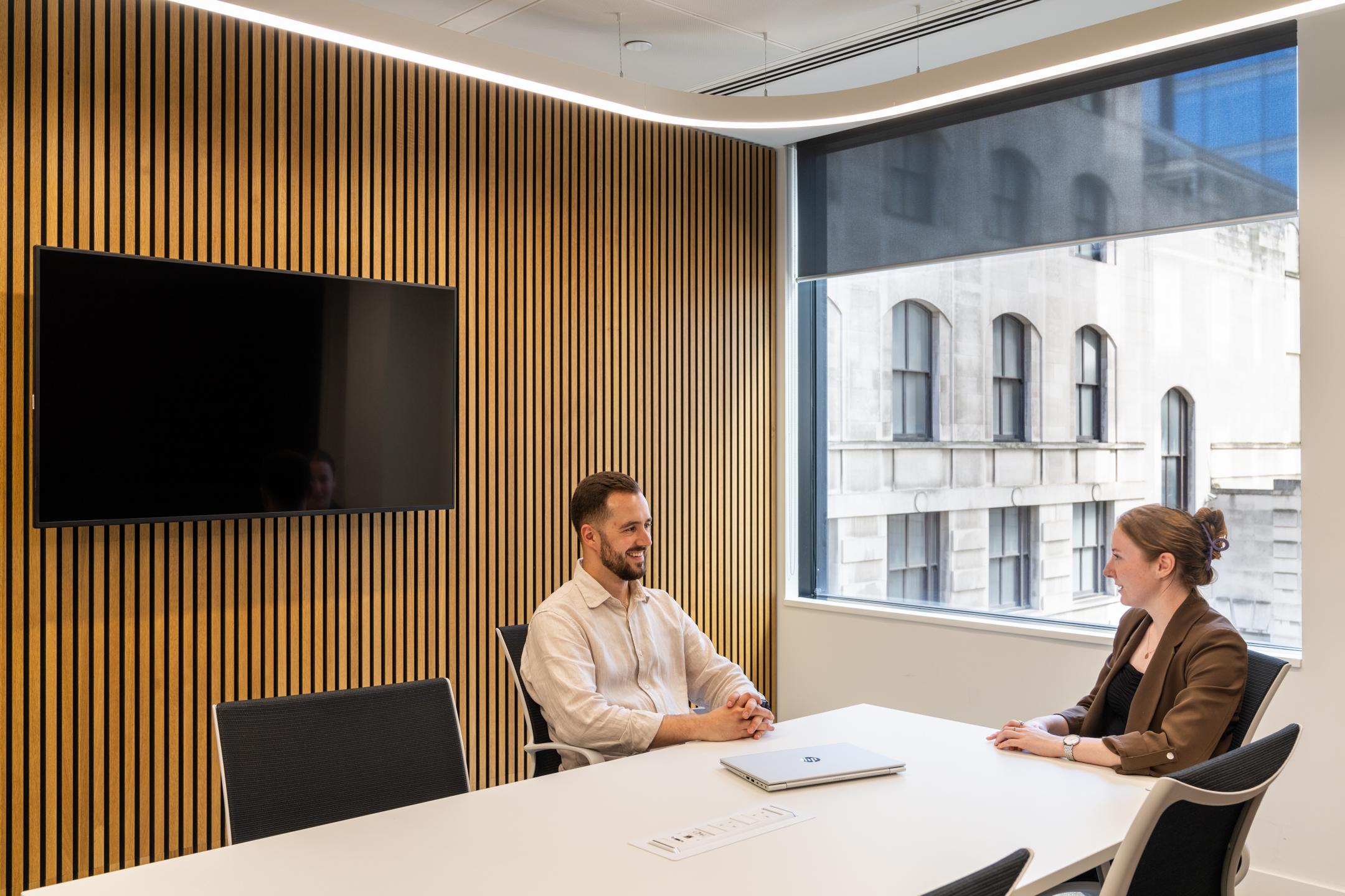 A small meeting room at DXC Technology with wooden slatted walls, a large window, and two people seated at a white table facing a wall-mounted screen.