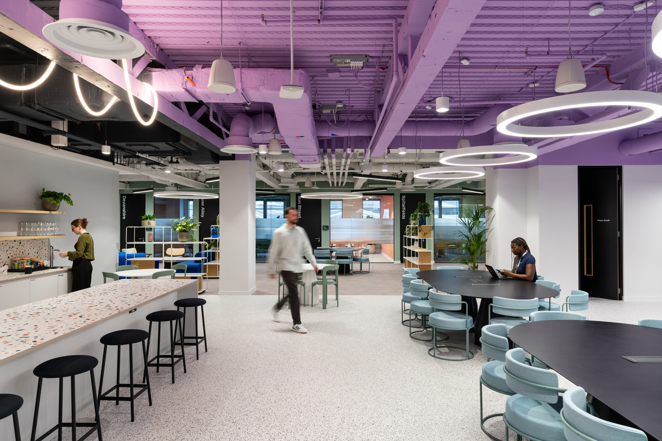 A bright, open teapoint and breakout area at Huckletree Bishopsgate, with bar stools lined up along a terrazzo-topped counter and circular light fixtures hanging from a purple ceiling.