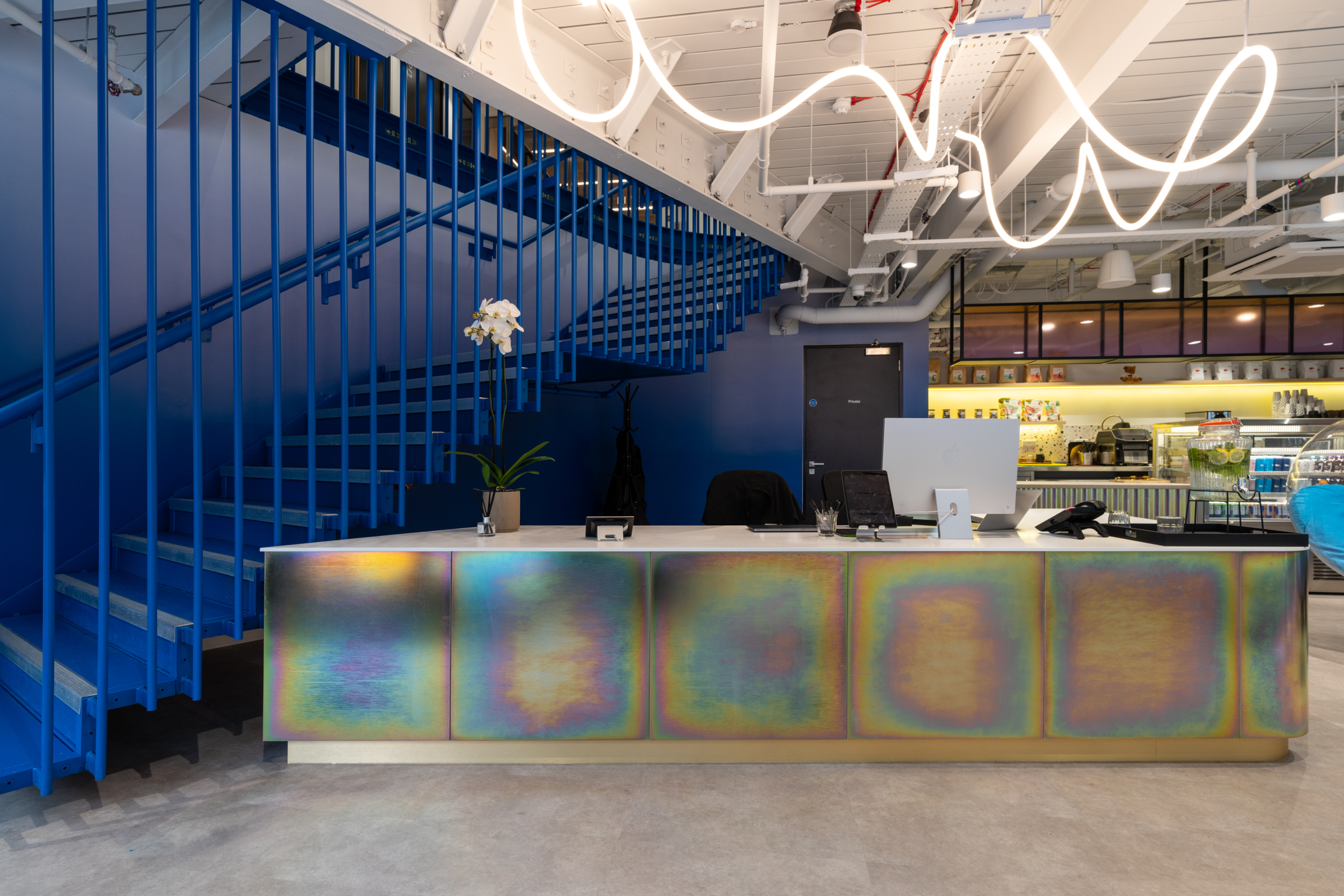 The reception area at Huckletree Bishopsgate has a colourful metallic desk, a blue staircase in the background, and bright industrial lighting overhead.