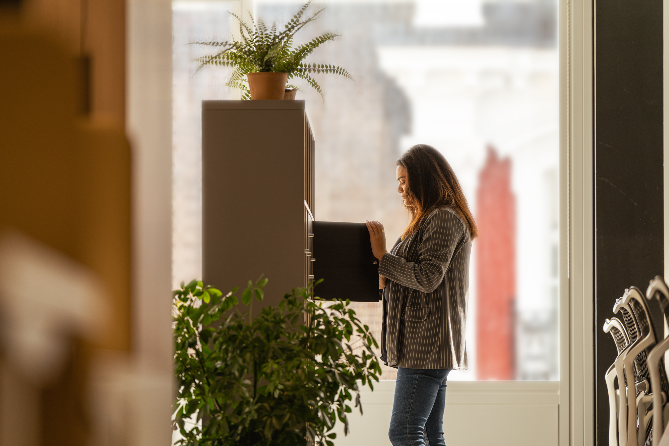 Woman browsing a folder in the Shionogi office, near potted plants and shelving.