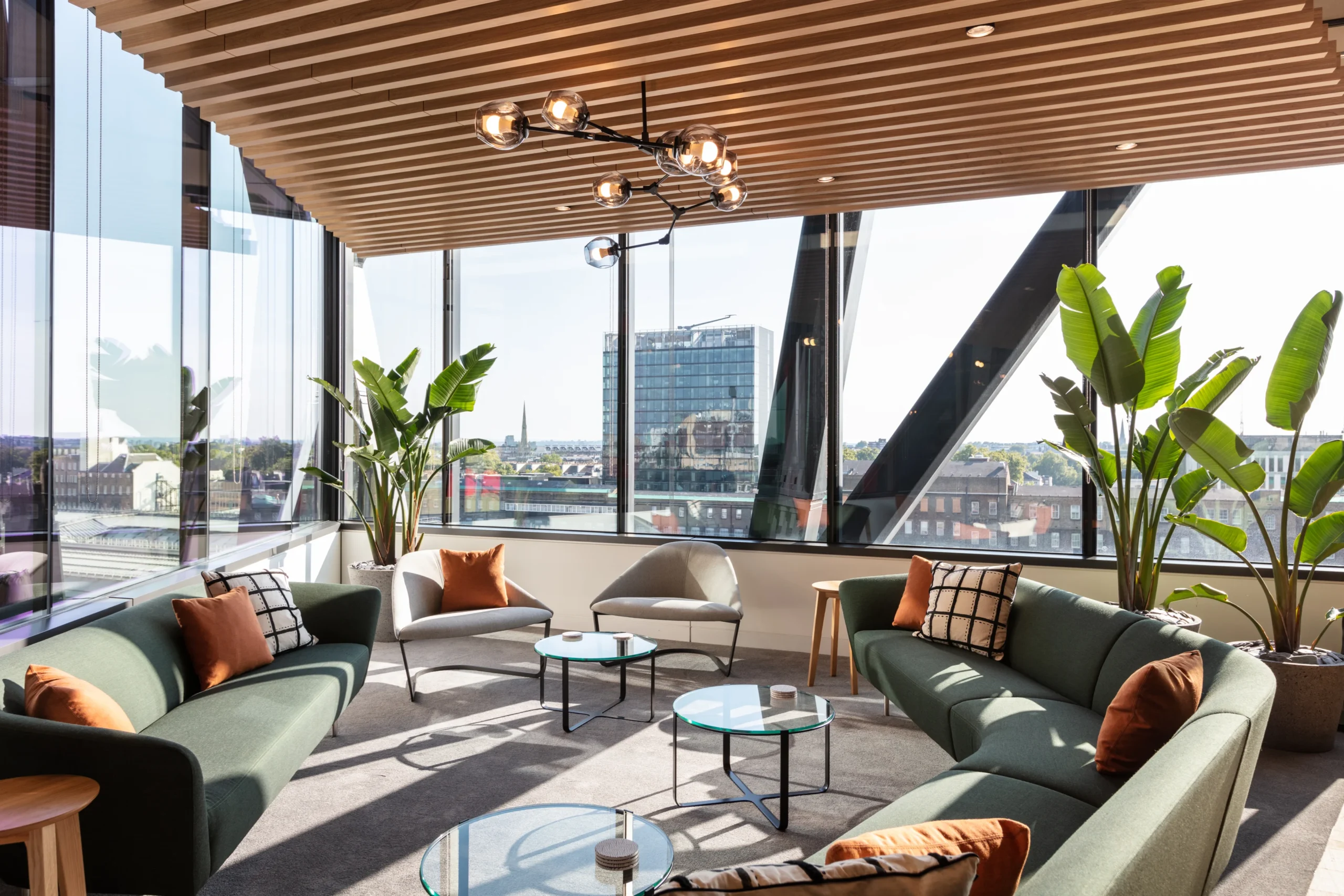 Alpha Fx’s breakout area with glass tables, soft seating, and large lush potted plants, facing a skyline glass view