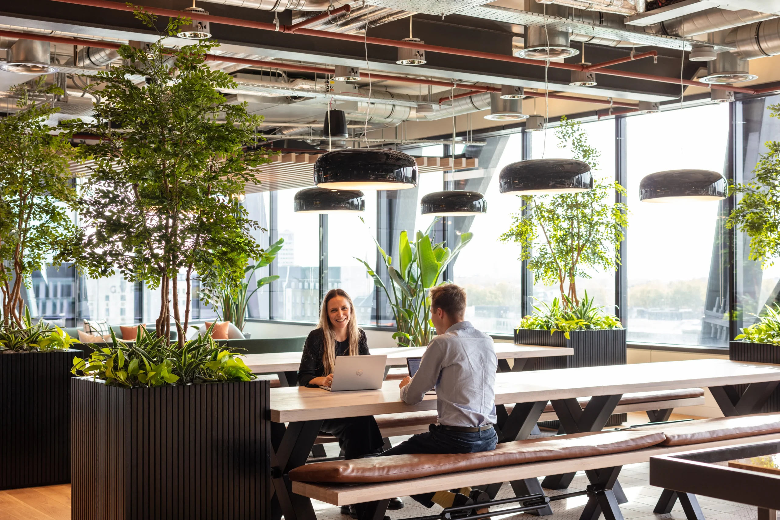 Alpha FX office designed by Modus Workspace, featuring a bright, open dining and collaboration space with long wooden tables, leather bench seating, and large indoor plants. Two people are seated at a table with laptops, working in a modern, nature-inspired environment with expansive windows providing natural light and city views.